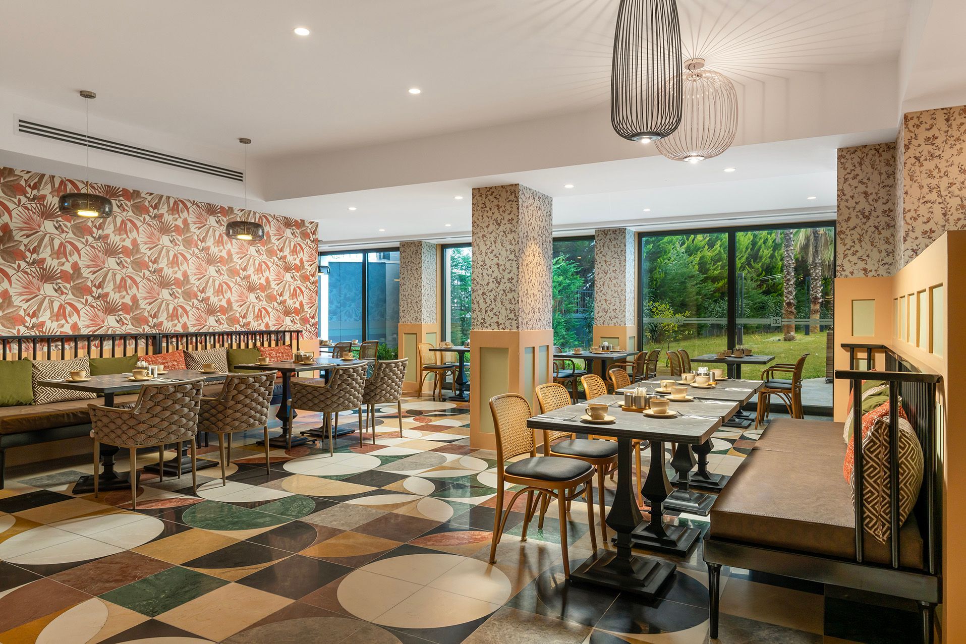 Bright, modern restaurant dining room with patterned carpet, tables set for service, and large windows overlooking greenery.