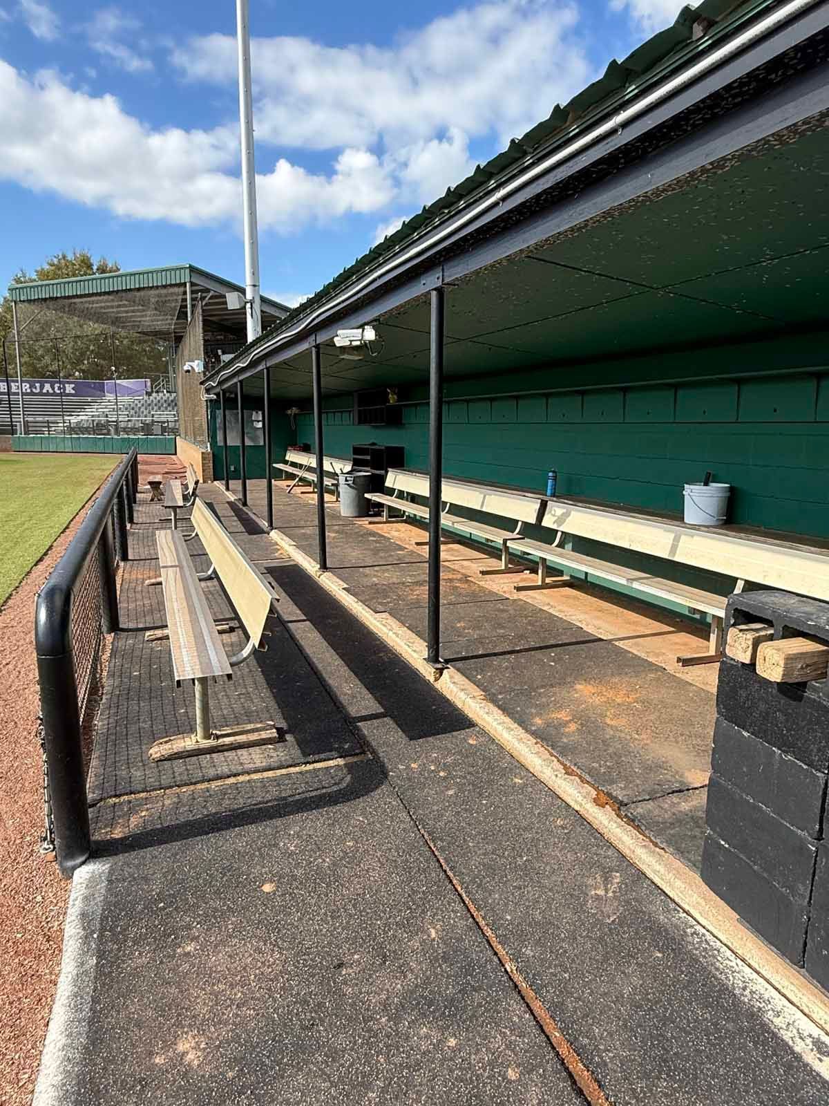 Baseball dugout with benches under a green roof. Black poles and a chain-link fence are visible.