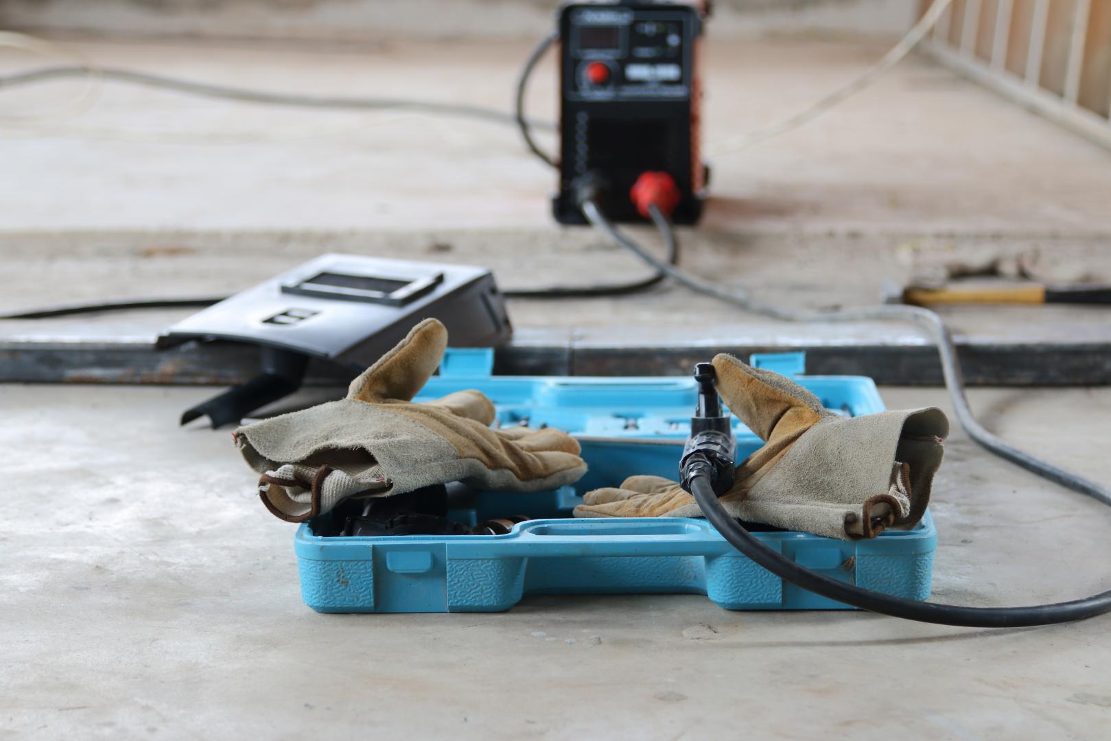 Des gants de soudage dans une boîte à outils bleue, un casque de soudage et une machine à souder sur un sol en béton.