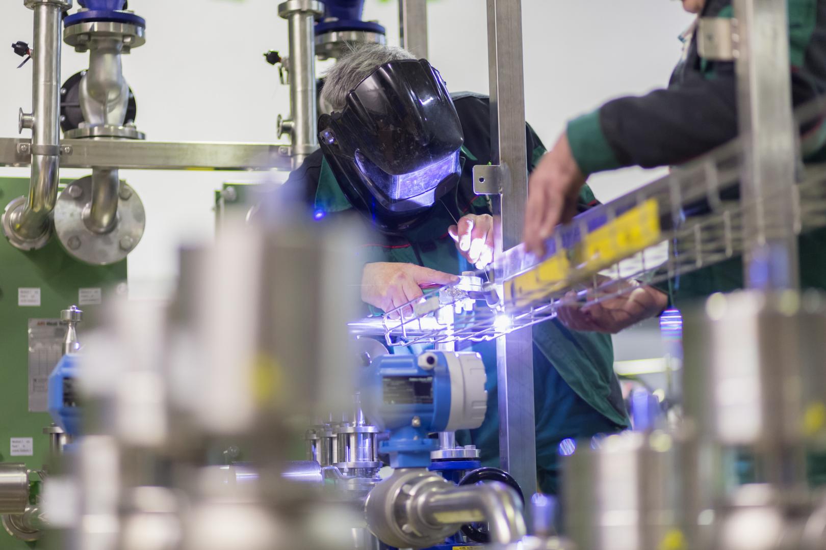 Une personne portant un masque de soudeur soude des tuyaux métalliques dans un environnement industriel.