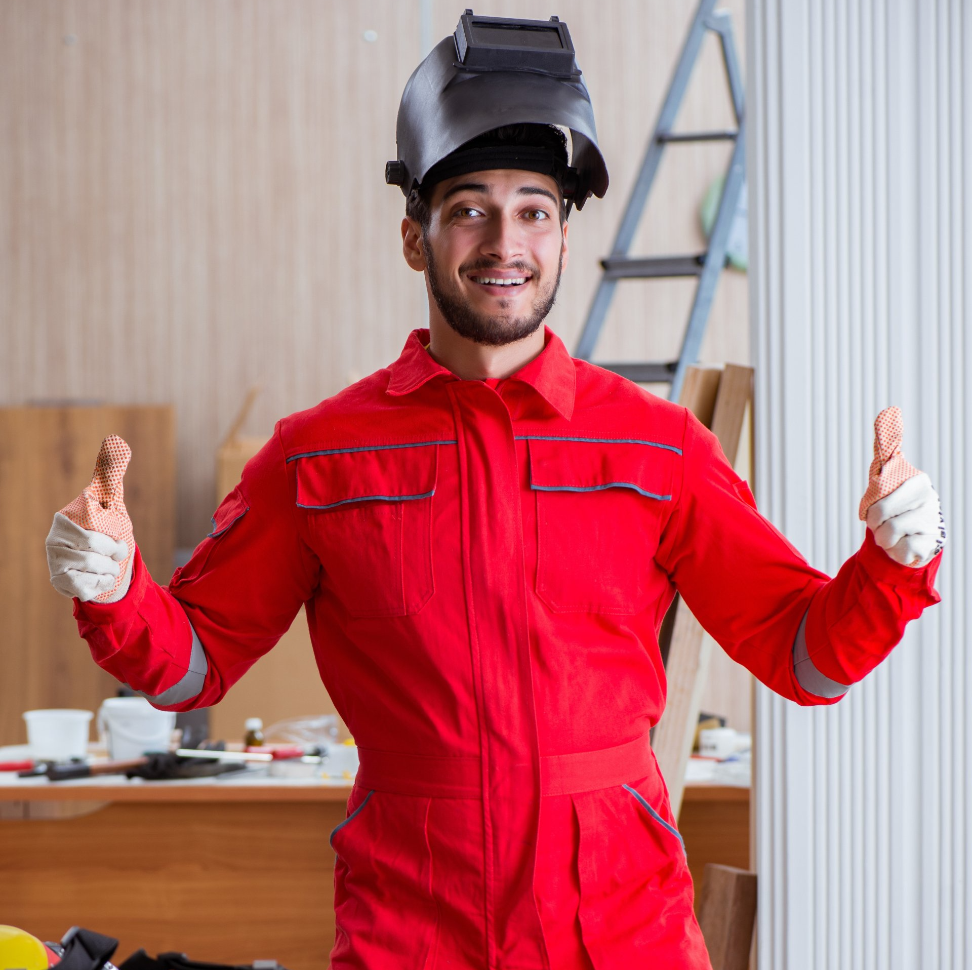 Un soudeur en combinaison rouge et casque lève le pouce dans un atelier.