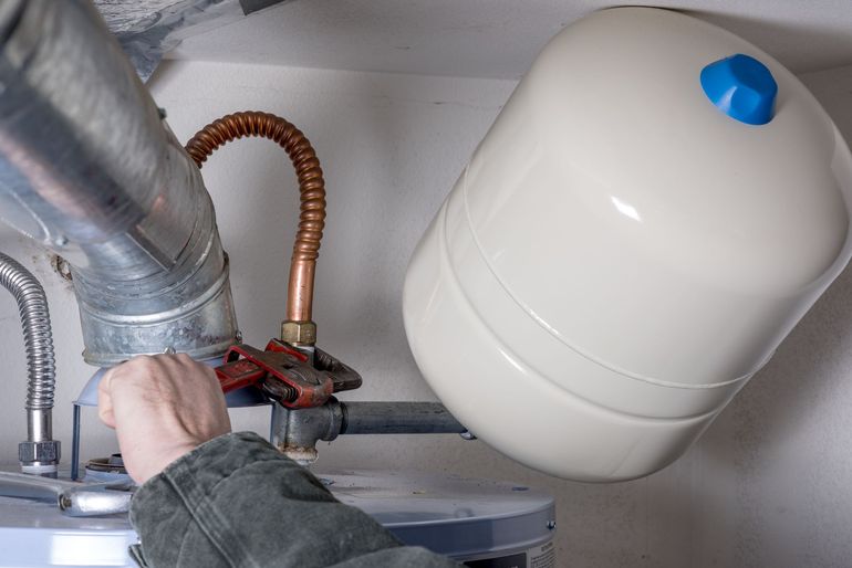 Hand connecting a copper pipe to a wall-mounted plumbing system beside a white expansion tank