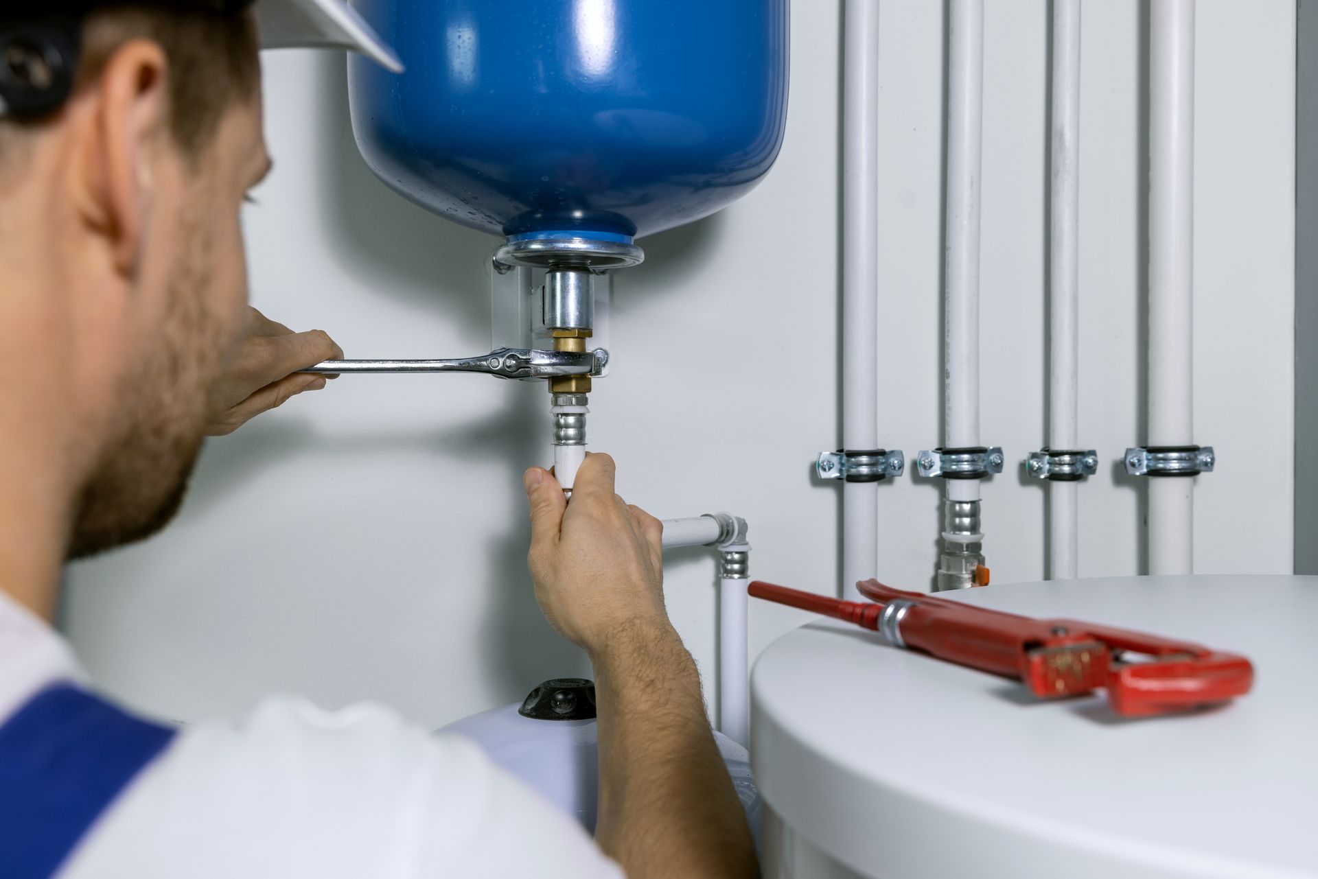 Plumber tightening a pipe under a blue water tank beside red pipe wrenches on a white wall.