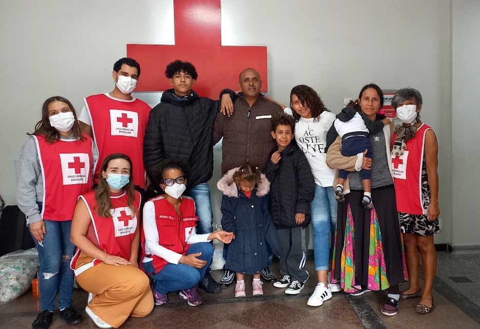 Um grupo de pessoas usando máscaras posa para uma foto em frente a uma cruz vermelha.