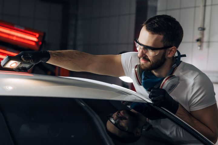 Un técnico con gafas de seguridad y respirador inspecciona la pintura de un coche blanco