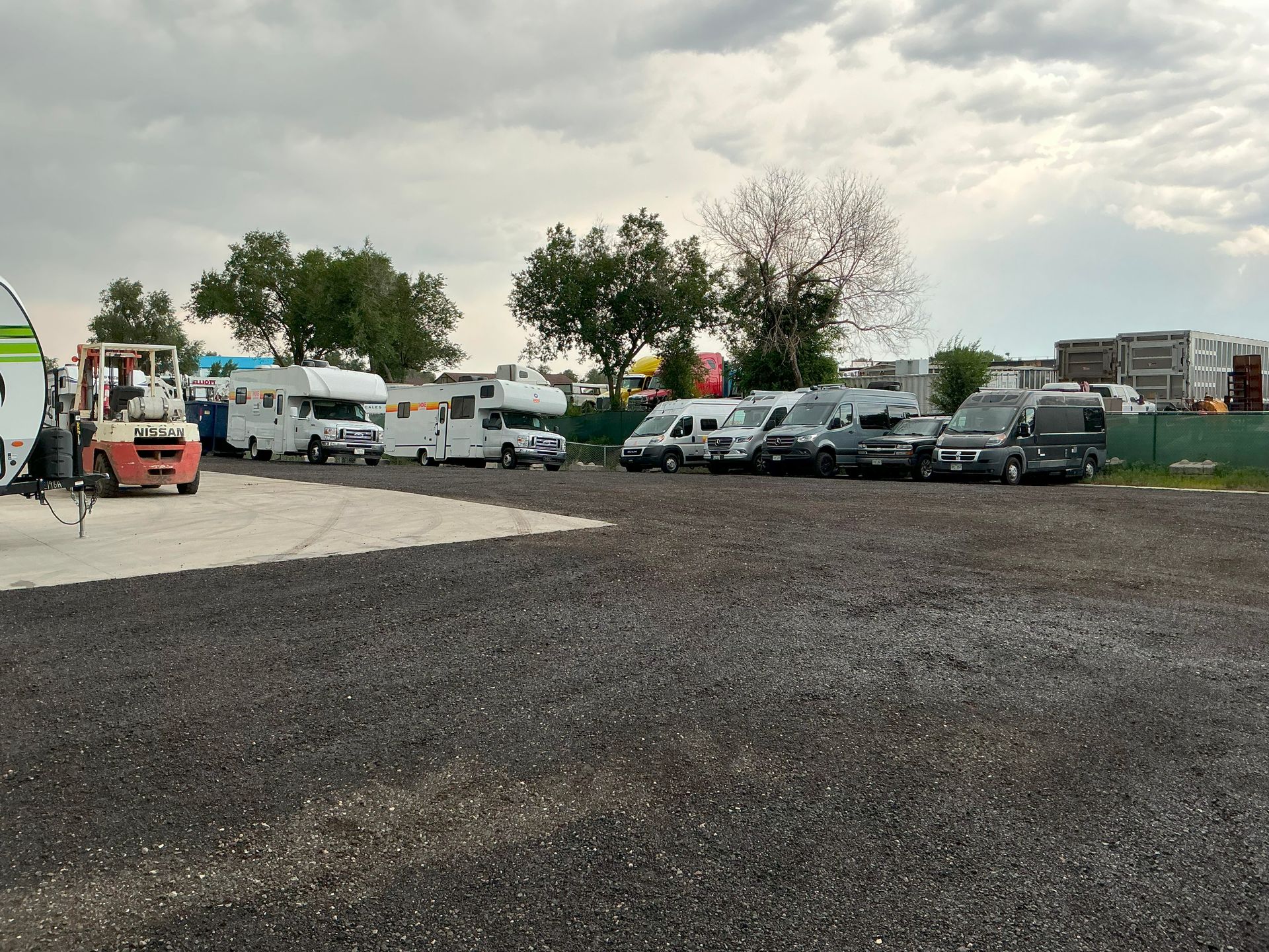 Vehicles parked on gravel; RVs, vans, and a forklift. Cloudy sky in the background.