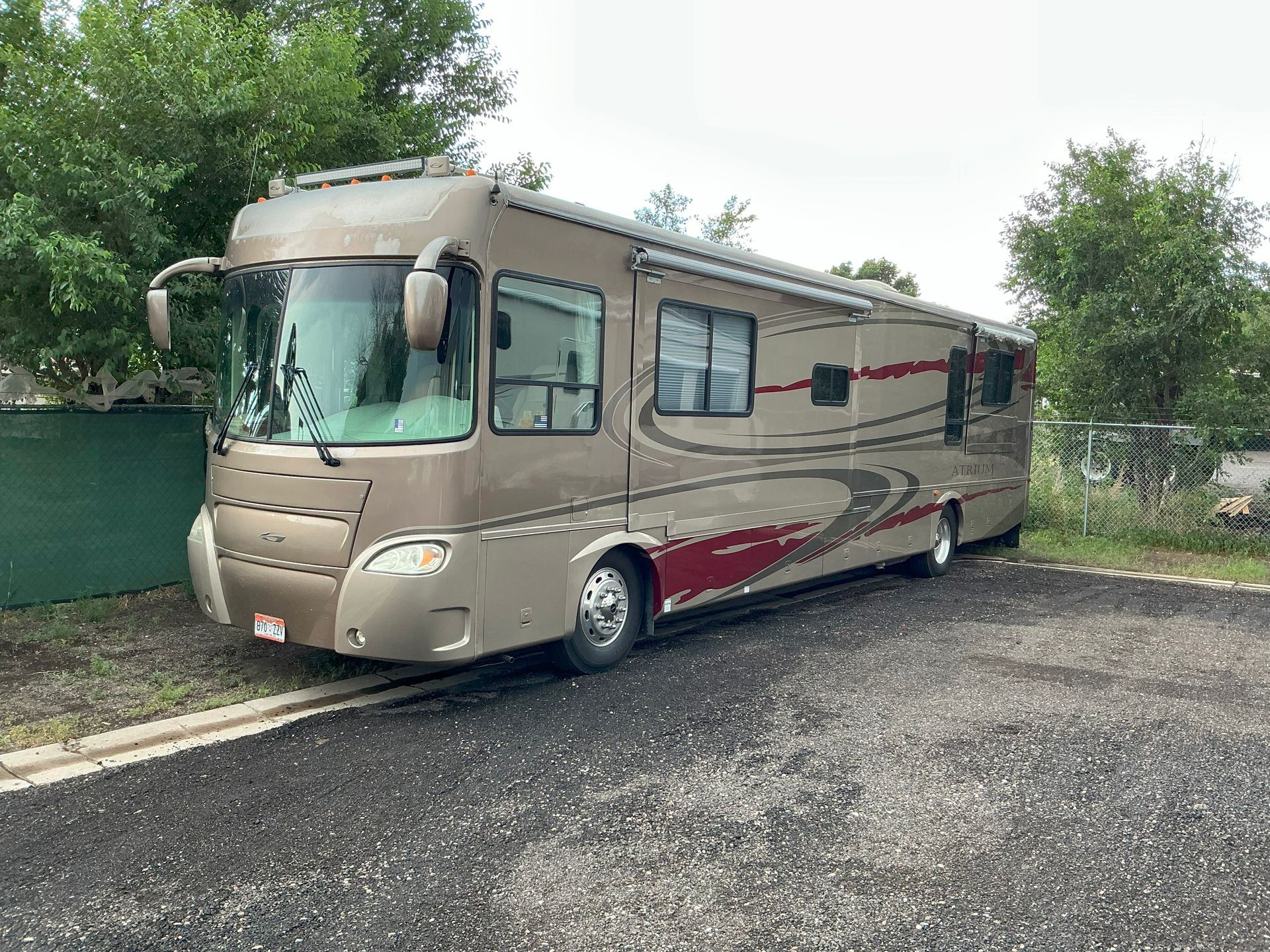 Tan and maroon RV parked on gravel next to a small building and some trees.