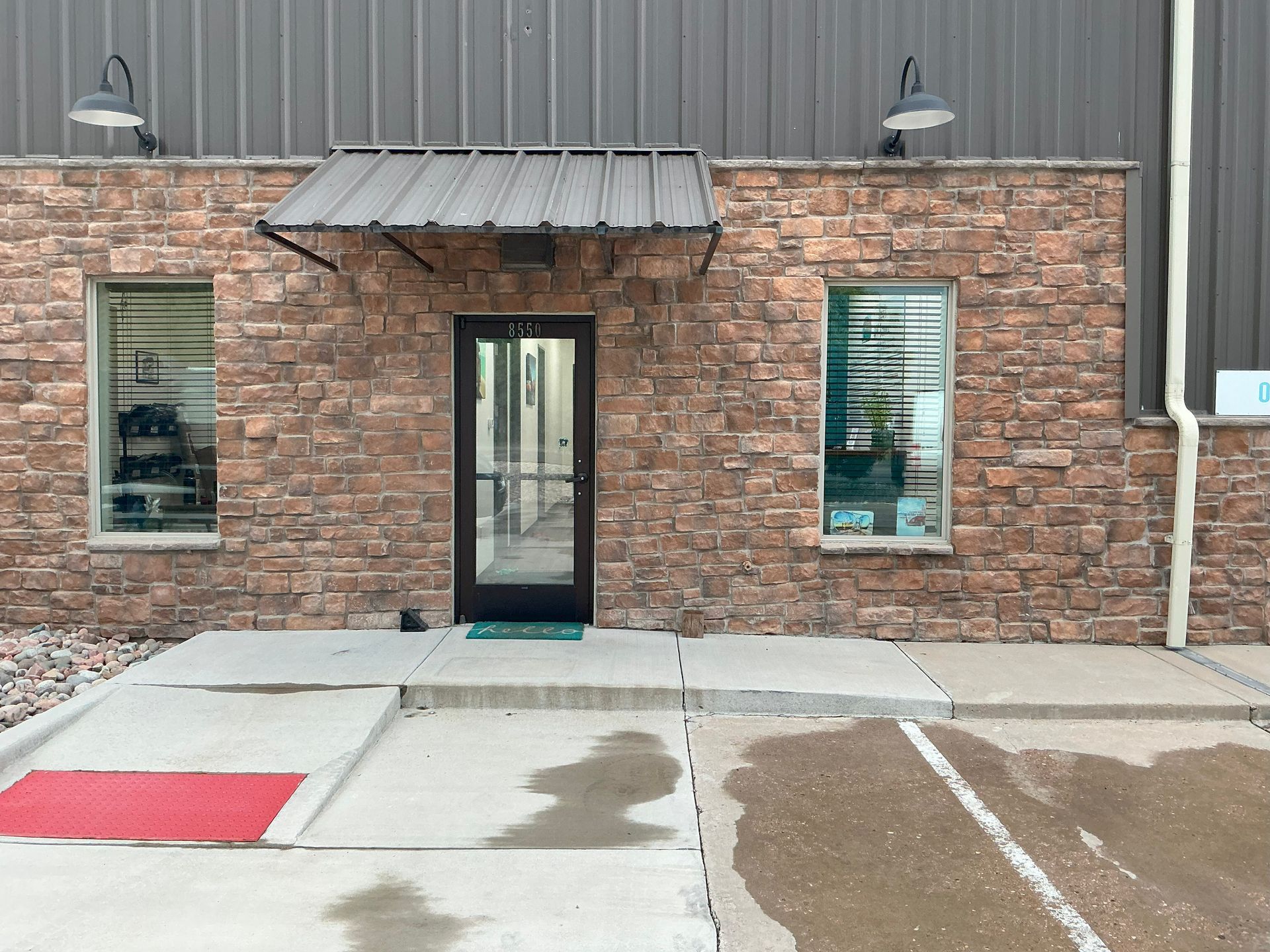Exterior of a building with brick facade, metal awning, glass door, two windows, and overhead lights.