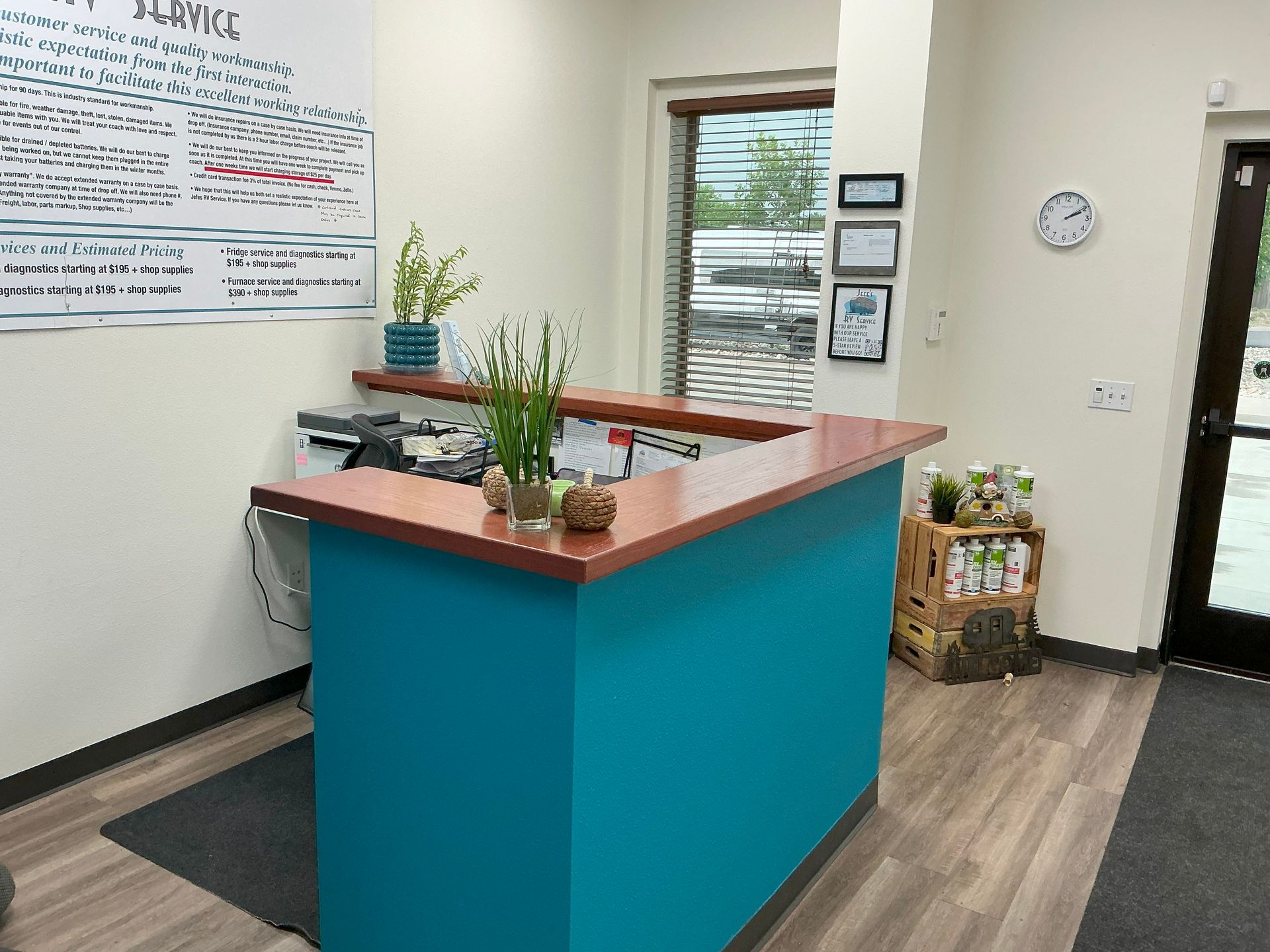 Teal reception desk with brown countertop, potted plants, and wooden flooring. Door and window in the background.
