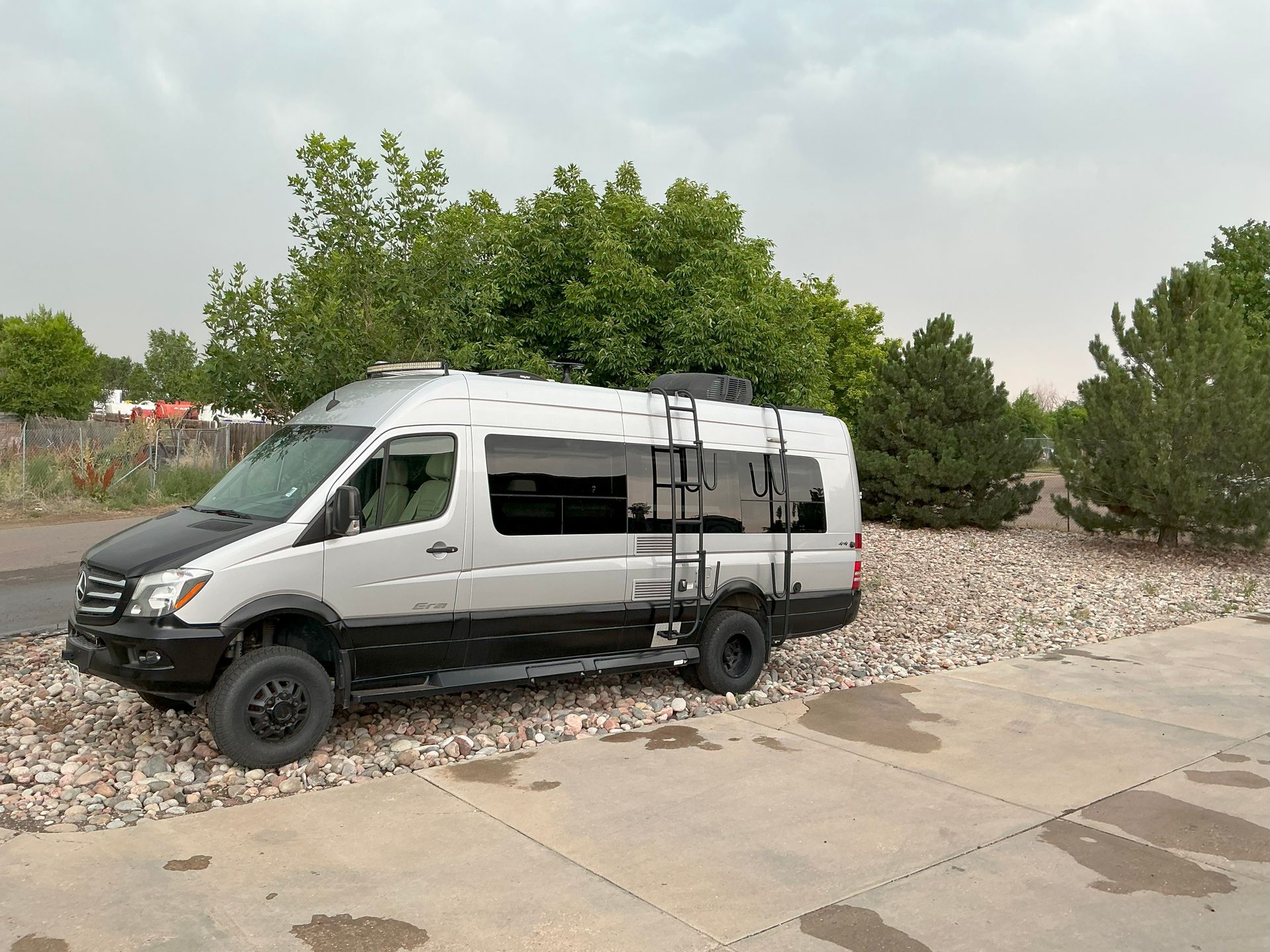 Silver and black camper van parked on gravel, with ladder, near trees and cloudy sky.