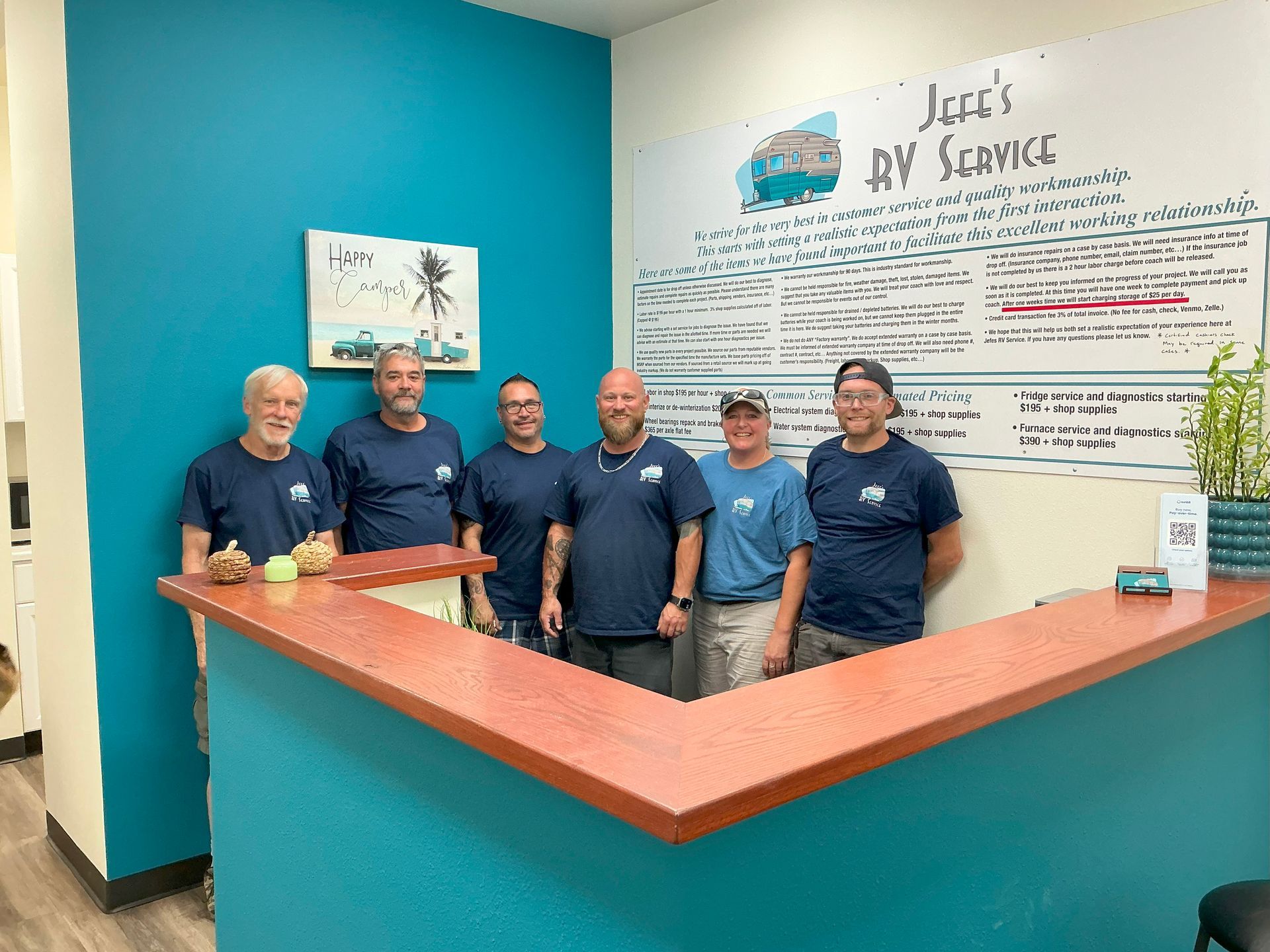 Six people in matching shirts at a reception desk. Teal walls, whiteboard, and a plant.