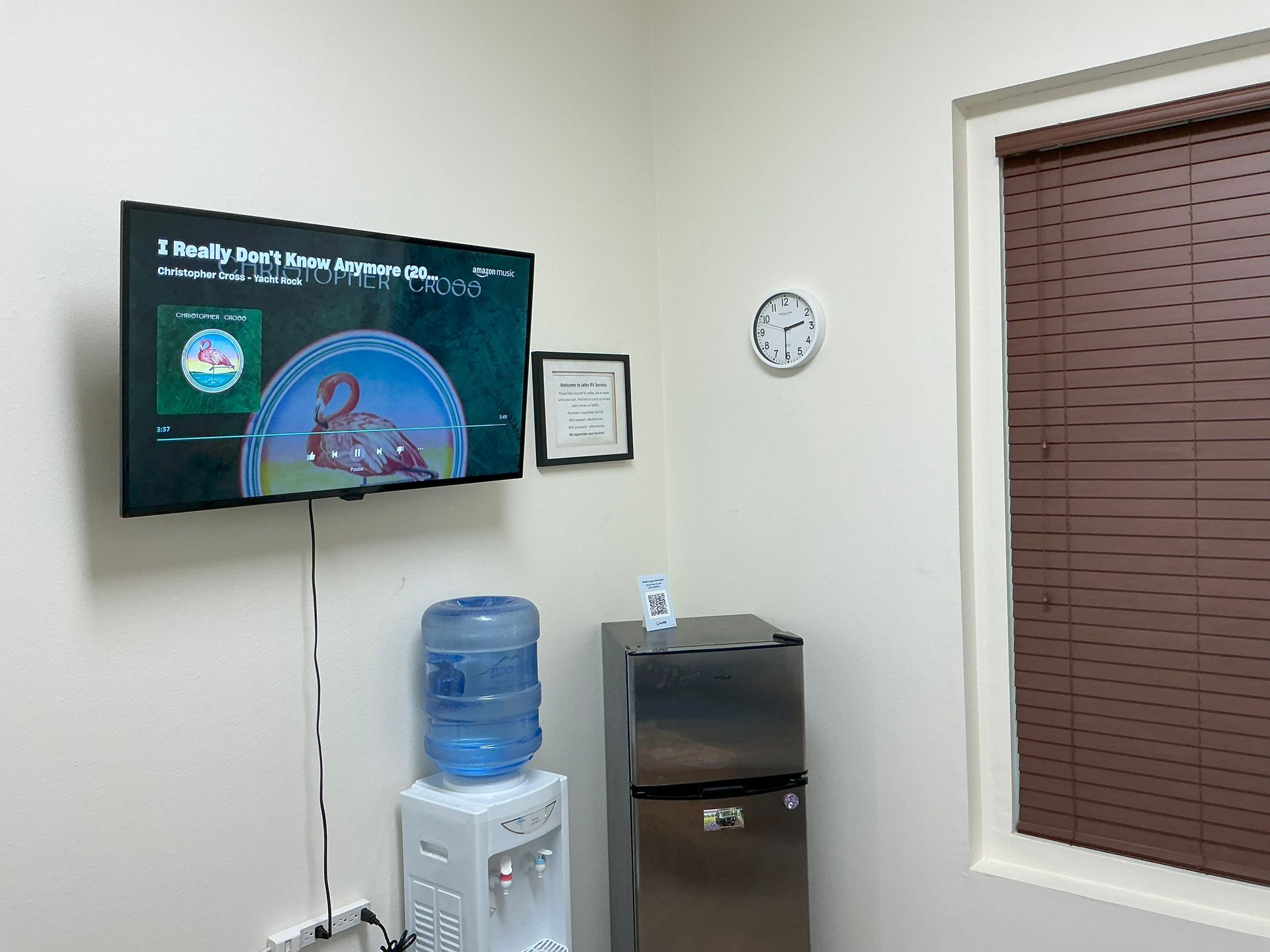 A small waiting room with a TV, water cooler, refrigerator, clock, and window with blinds.