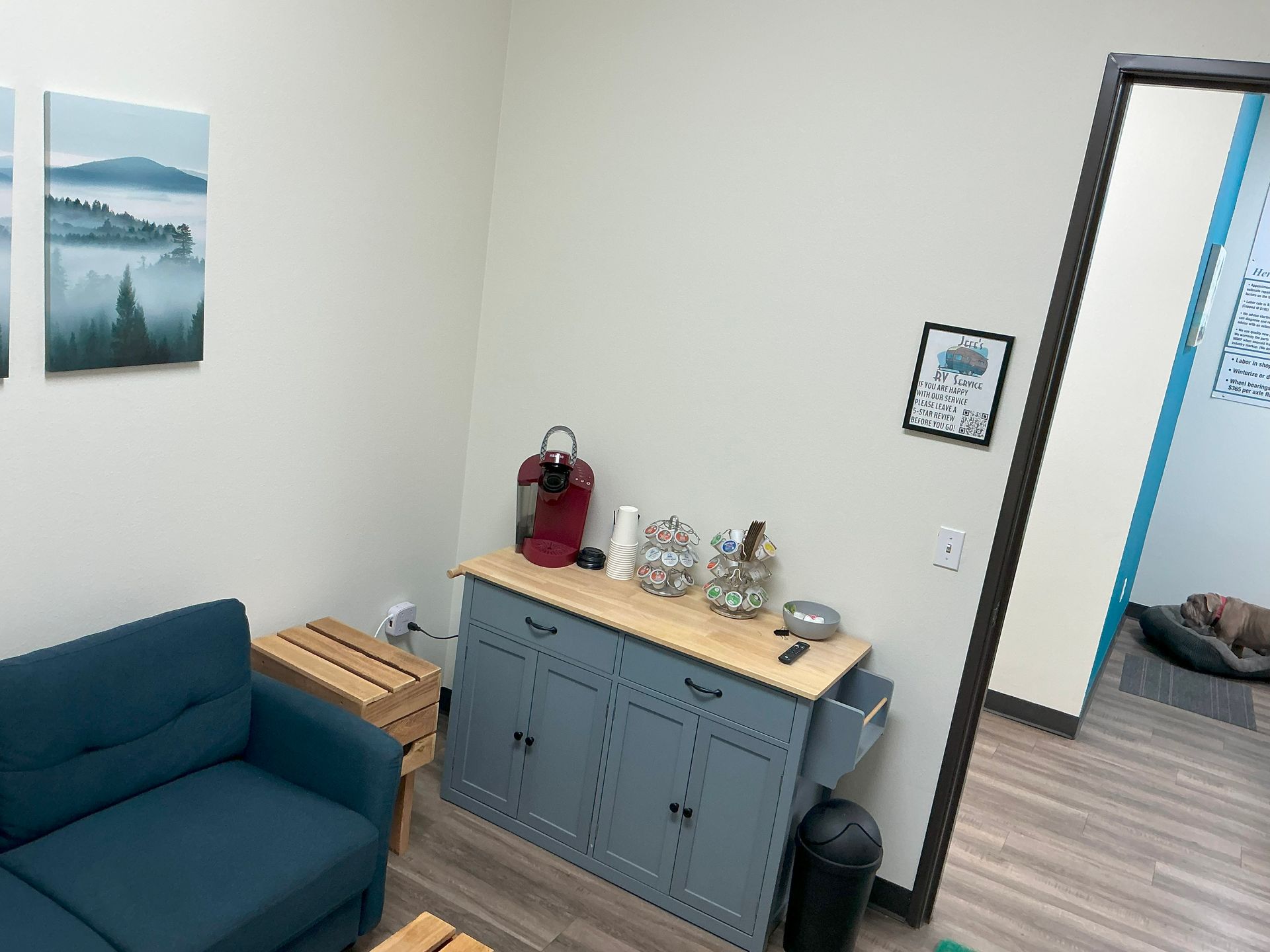 A room with a blue couch, coffee station, and artwork. A dog sleeps in the doorway.