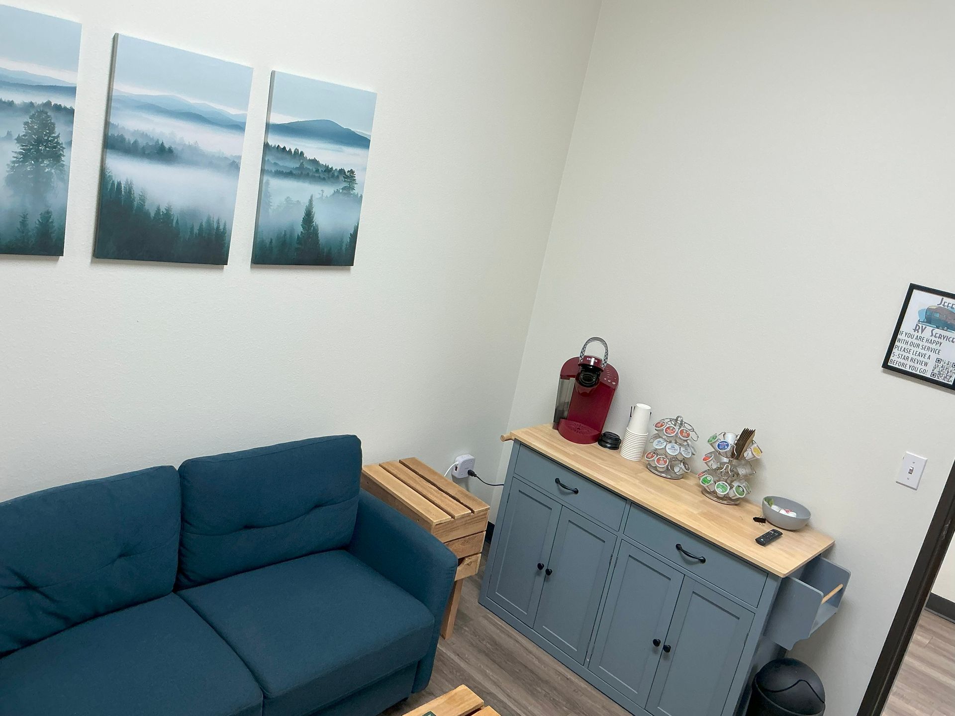 A teal couch in a waiting area with artwork, a cabinet with snacks, and a coffee maker.
