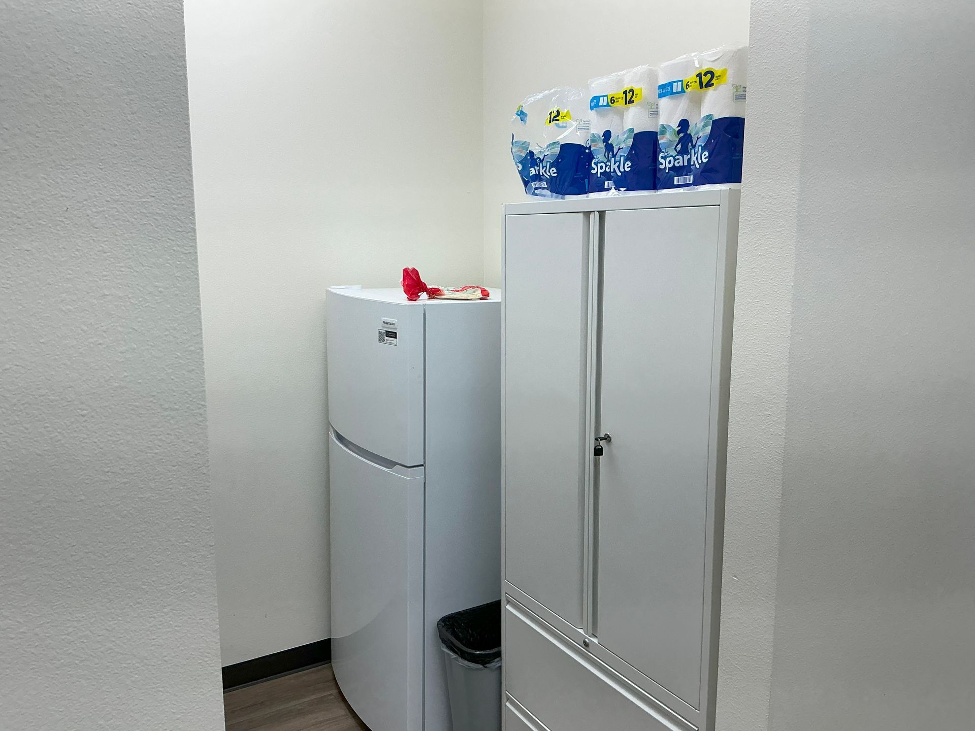 Refrigerator, white cabinet, and trash can in a small, white-walled room; toilet paper rolls on top of the cabinet.