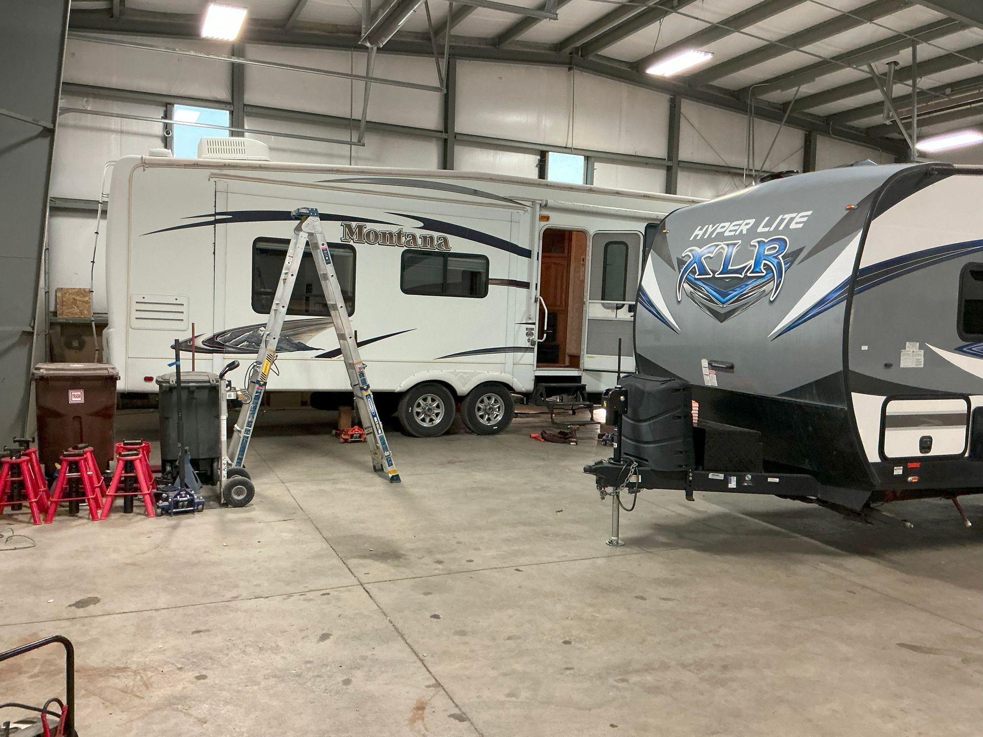 Two RVs inside a large garage. One is white, being worked on with a ladder. The other is gray, in the foreground.