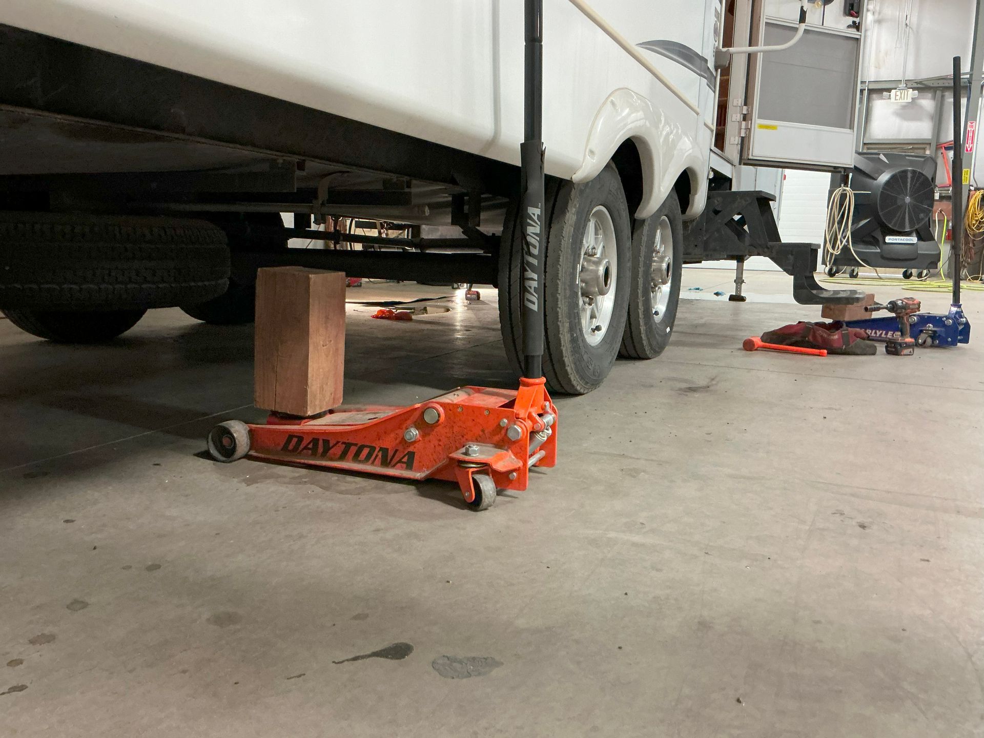 An RV is jacked up in a garage. An orange jack and a wooden block are used to lift it.