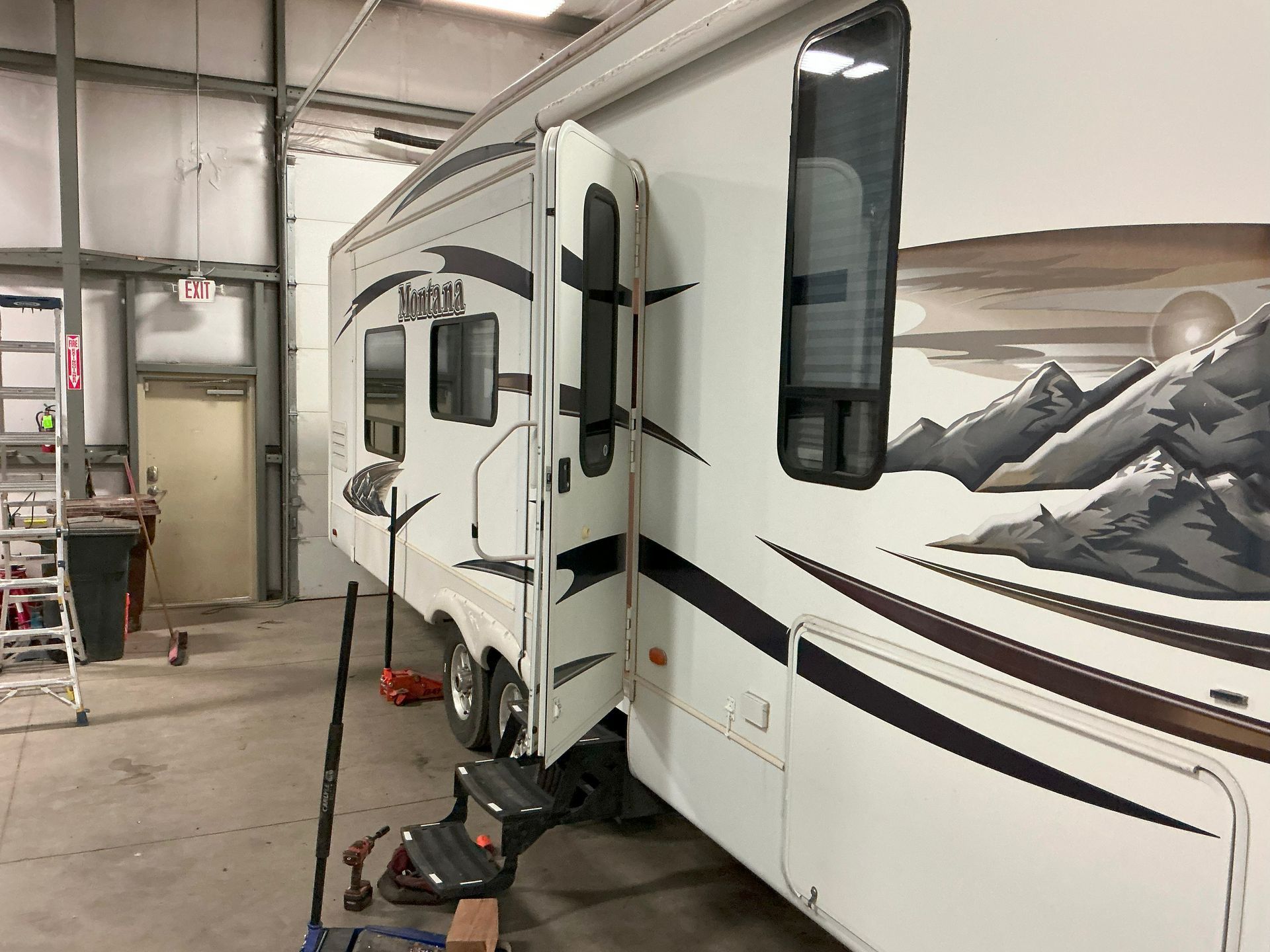 RV trailer with slide-out extended in a garage, showcasing mountain artwork on its side.