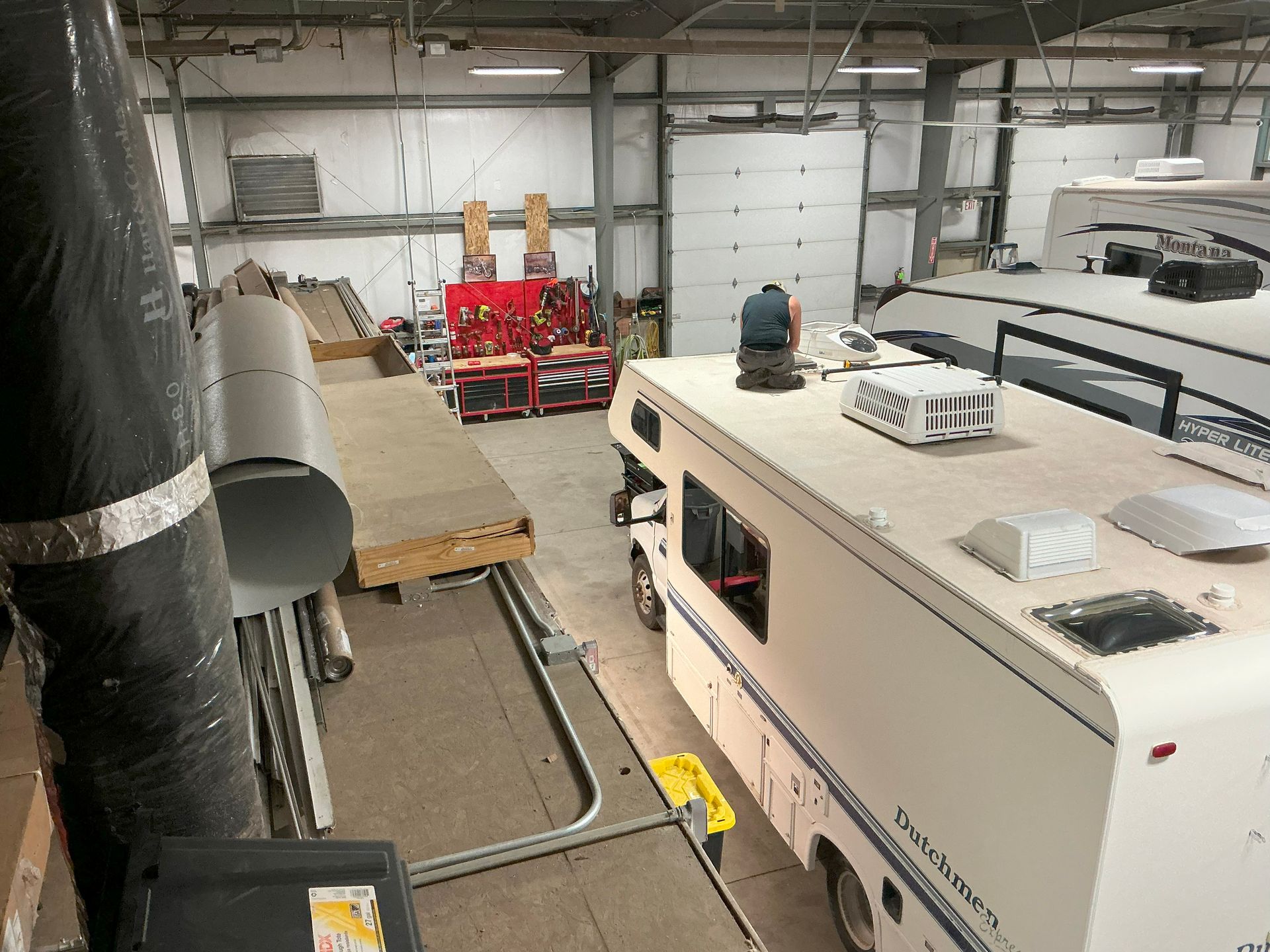 Inside a large workshop, an RV is being worked on, with tools and a person visible on the roof.