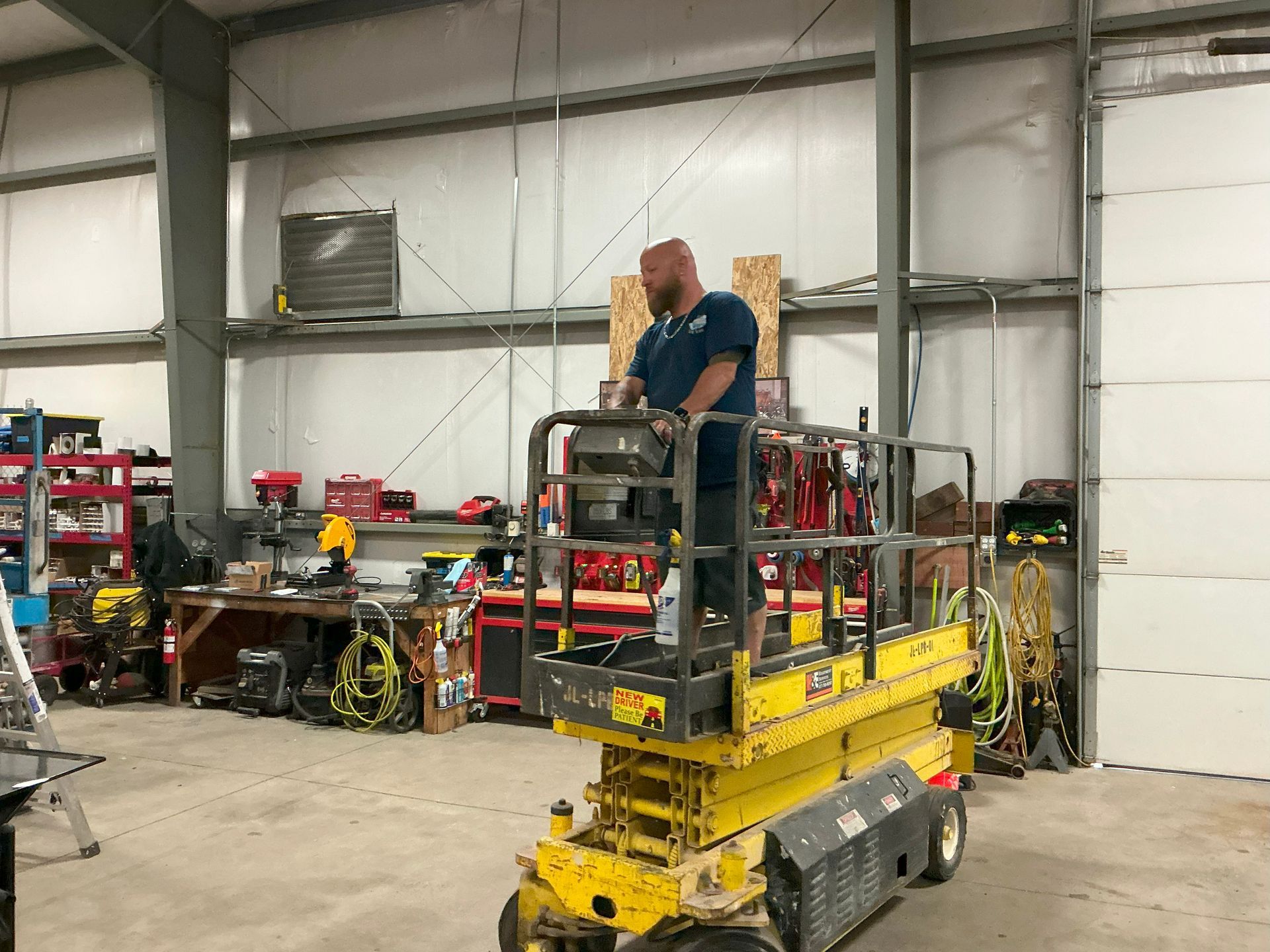 Man on yellow lift inside a garage, working on overhead fixtures. Tools and equipment visible.