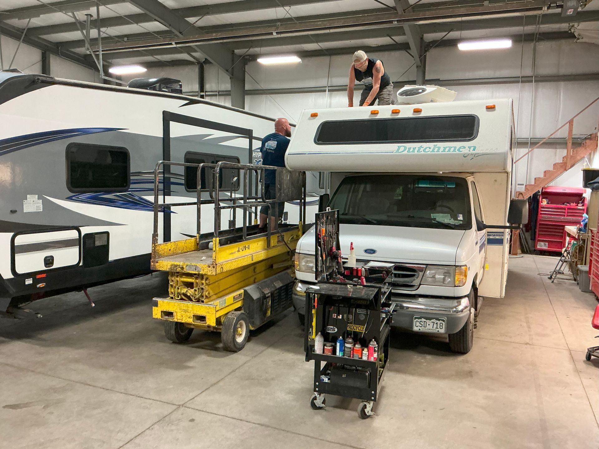Two people work on a RV roof in a garage. One on a lift, one on the roof. RV and tool cart nearby.
