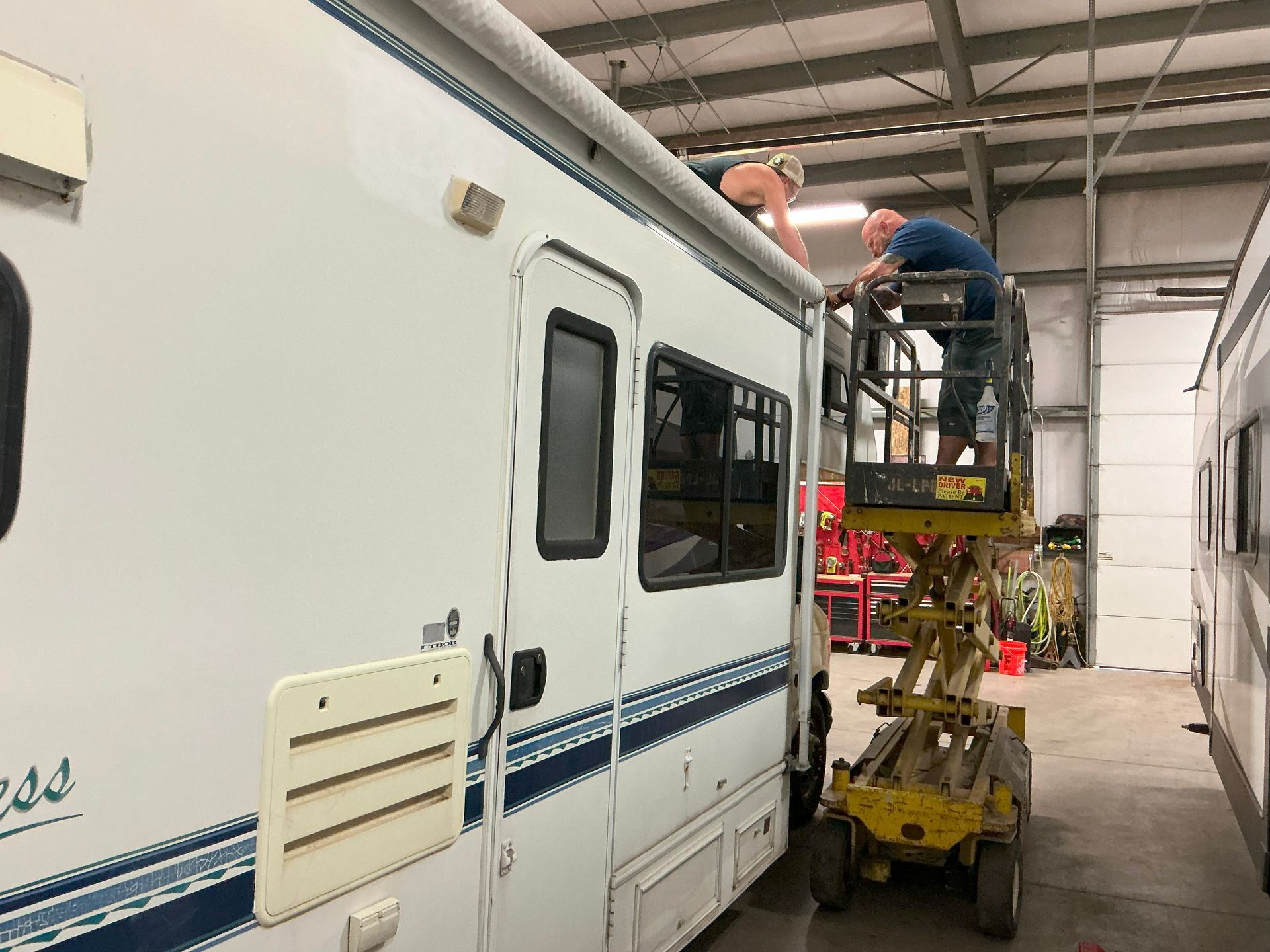 Two people working on the awning of a white RV, one on a lift, inside a garage.