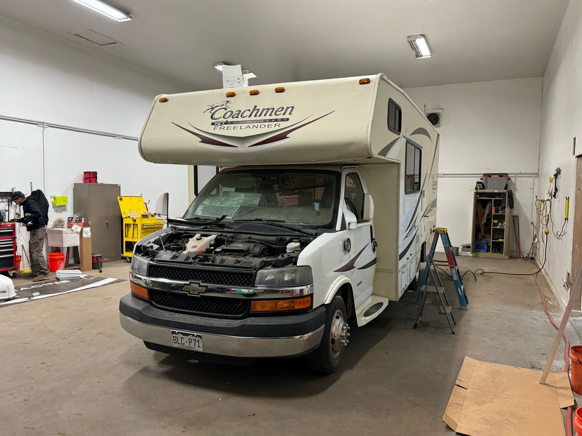 RV with open hood inside a garage. White and tan vehicle. Person working nearby.