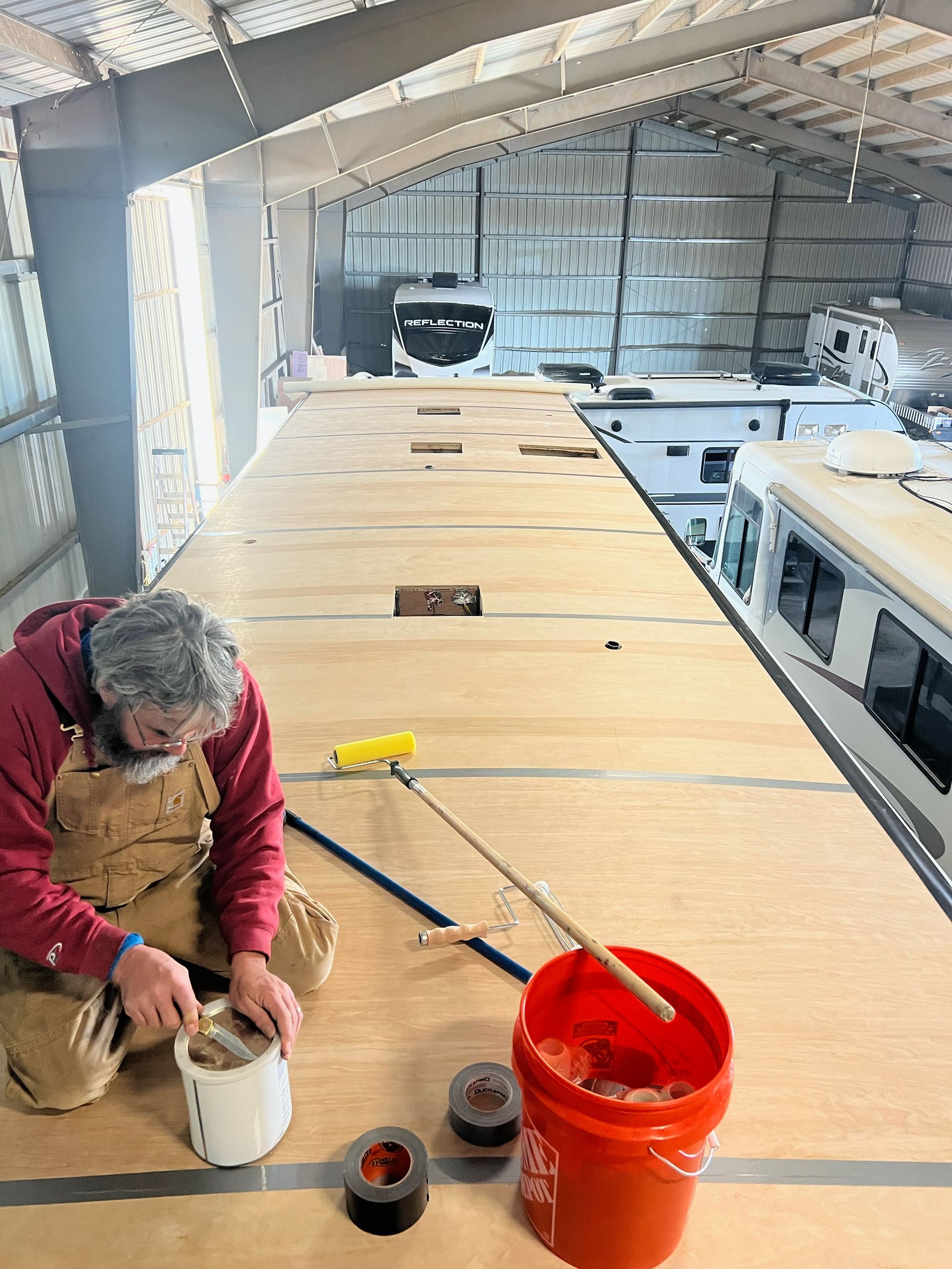 A person repairs a camper roof inside a large warehouse. They work near a bucket and tools.