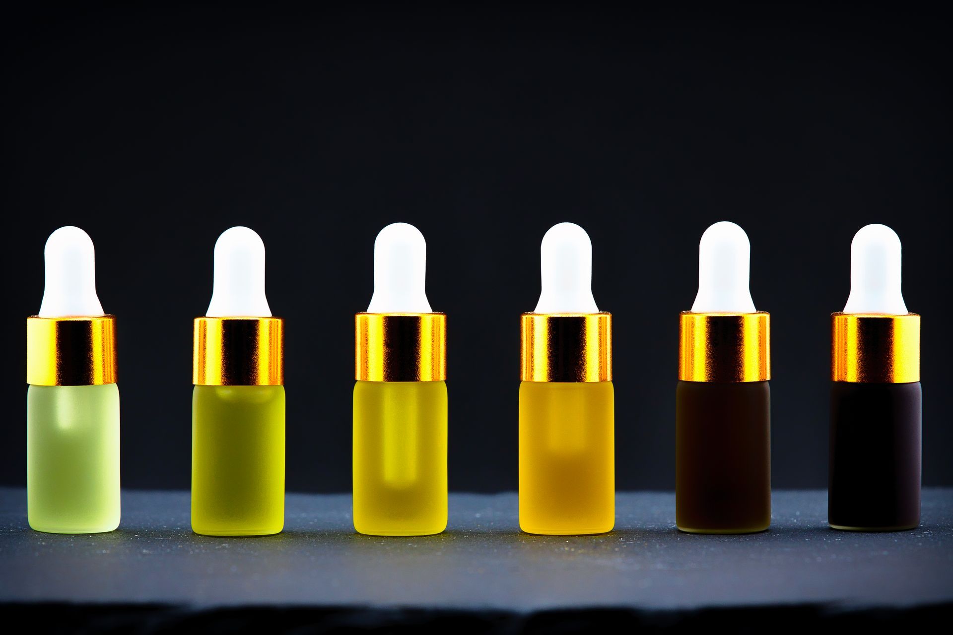 Six small dropper bottles with white caps in a row, showing a green-to-black color gradient.