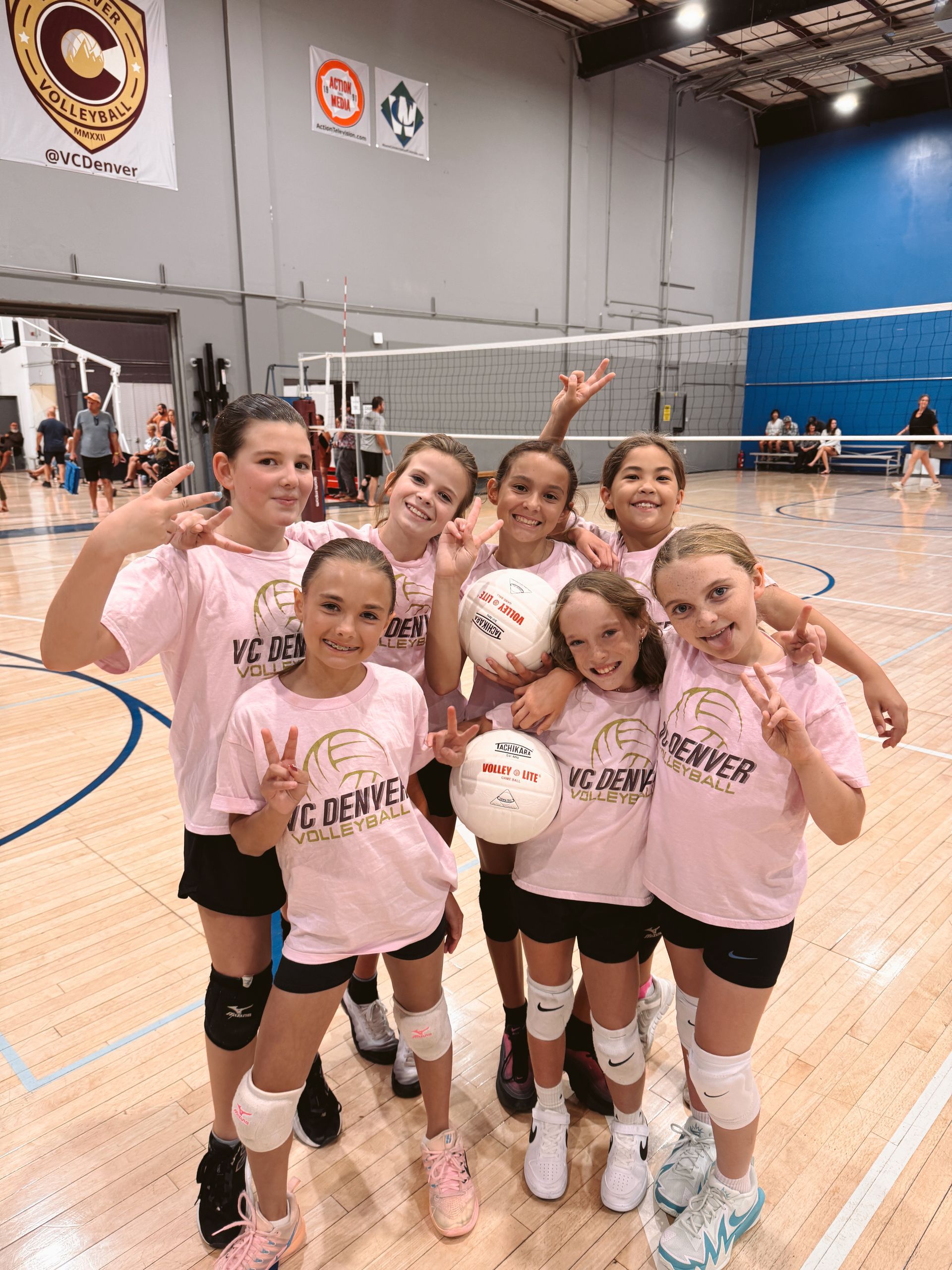 Girls, some carrying others piggyback style, pose with volleyballs in a gym, smiling.
