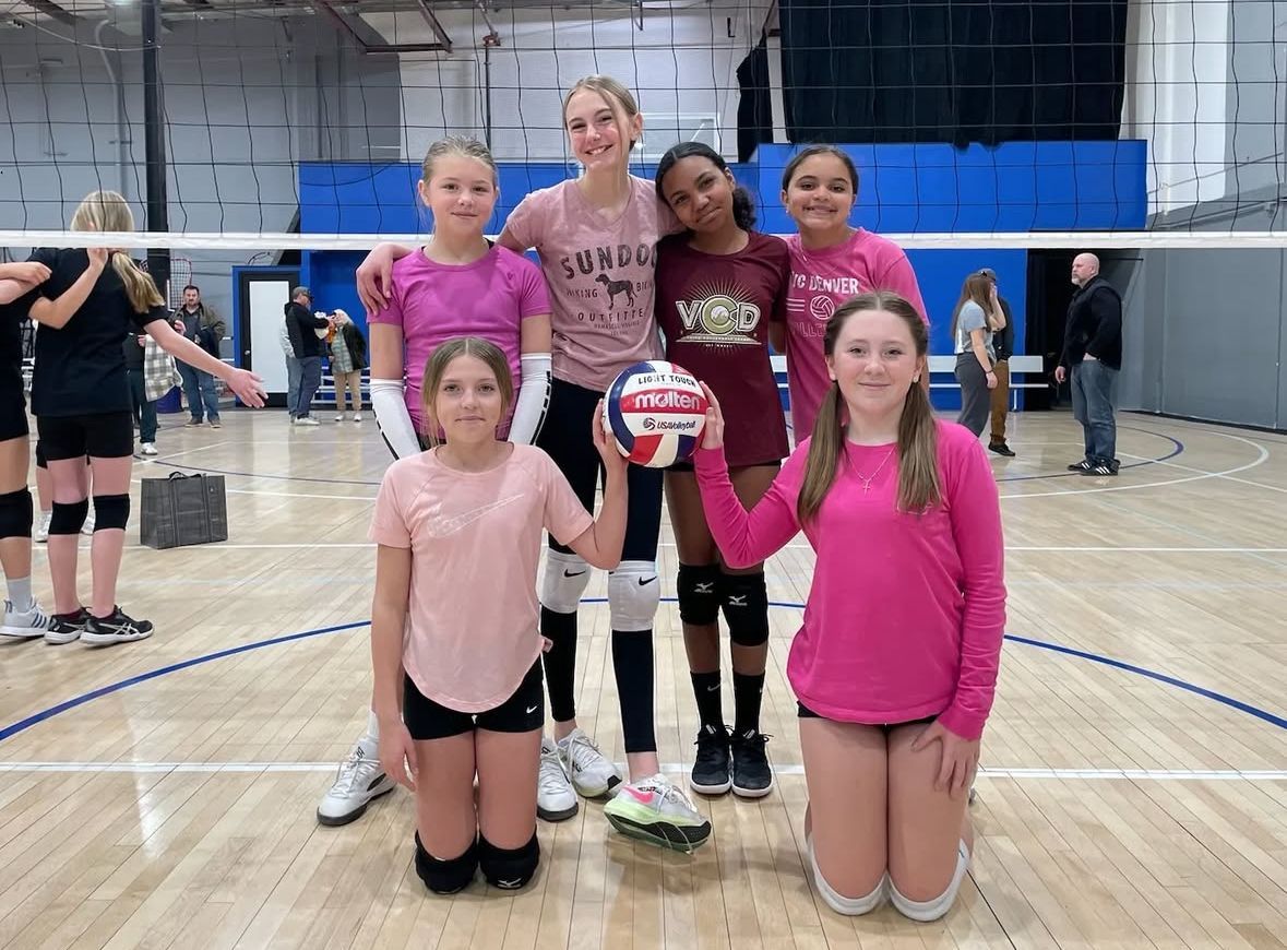 Girls volleyball team posing with ball in gym. Mostly pink shirts.