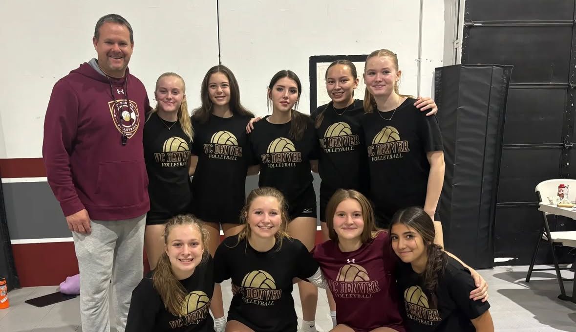 Group of people posing indoors in matching black shirts beside stacked speakers and a maroon-clad man.