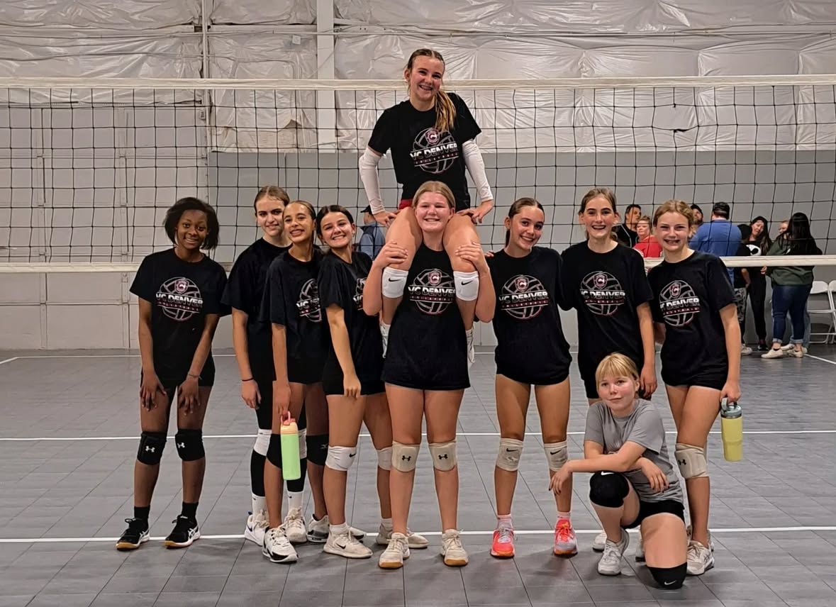 Volleyball team posing in a gym, one player on another’s shoulders, with a trophy in front