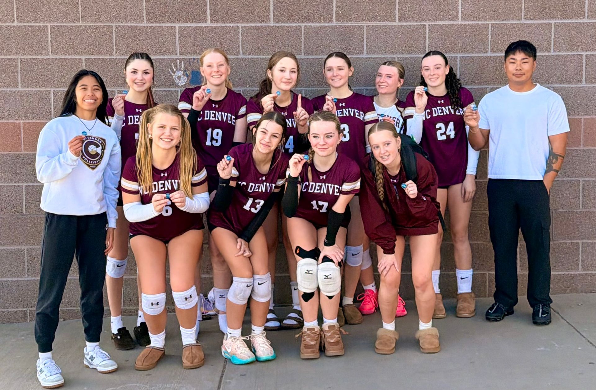 Volleyball team posing in maroon uniforms with coaches in front of a brick wall.