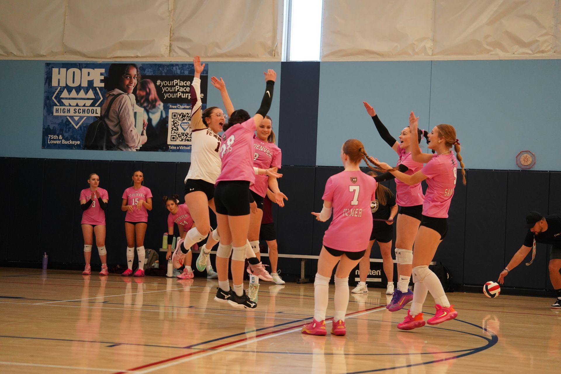 Volleyball players in pink jerseys celebrate with a jump on an indoor court