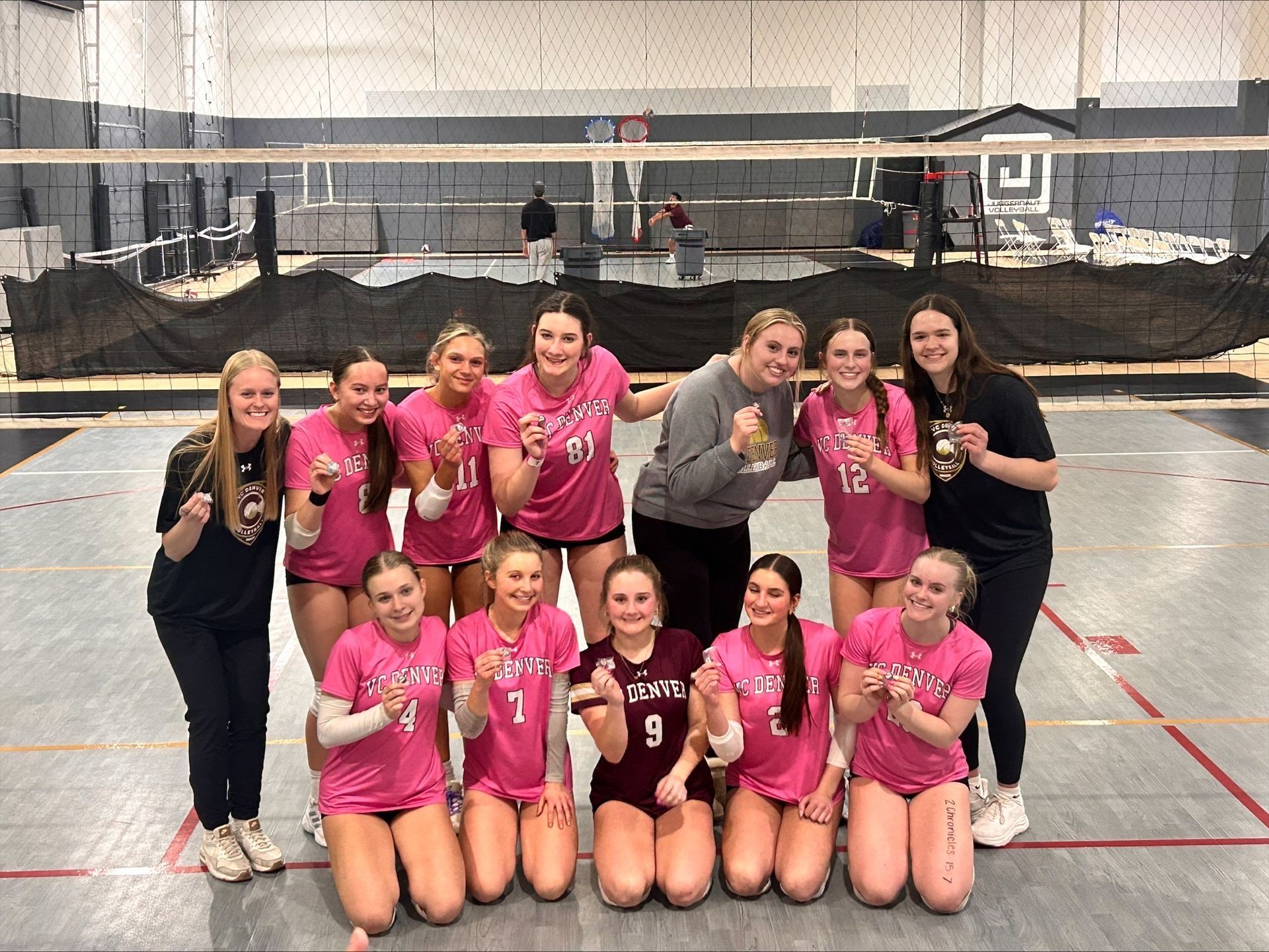 Volleyball team posing indoors on a court, many in pink jerseys, smiling after a match