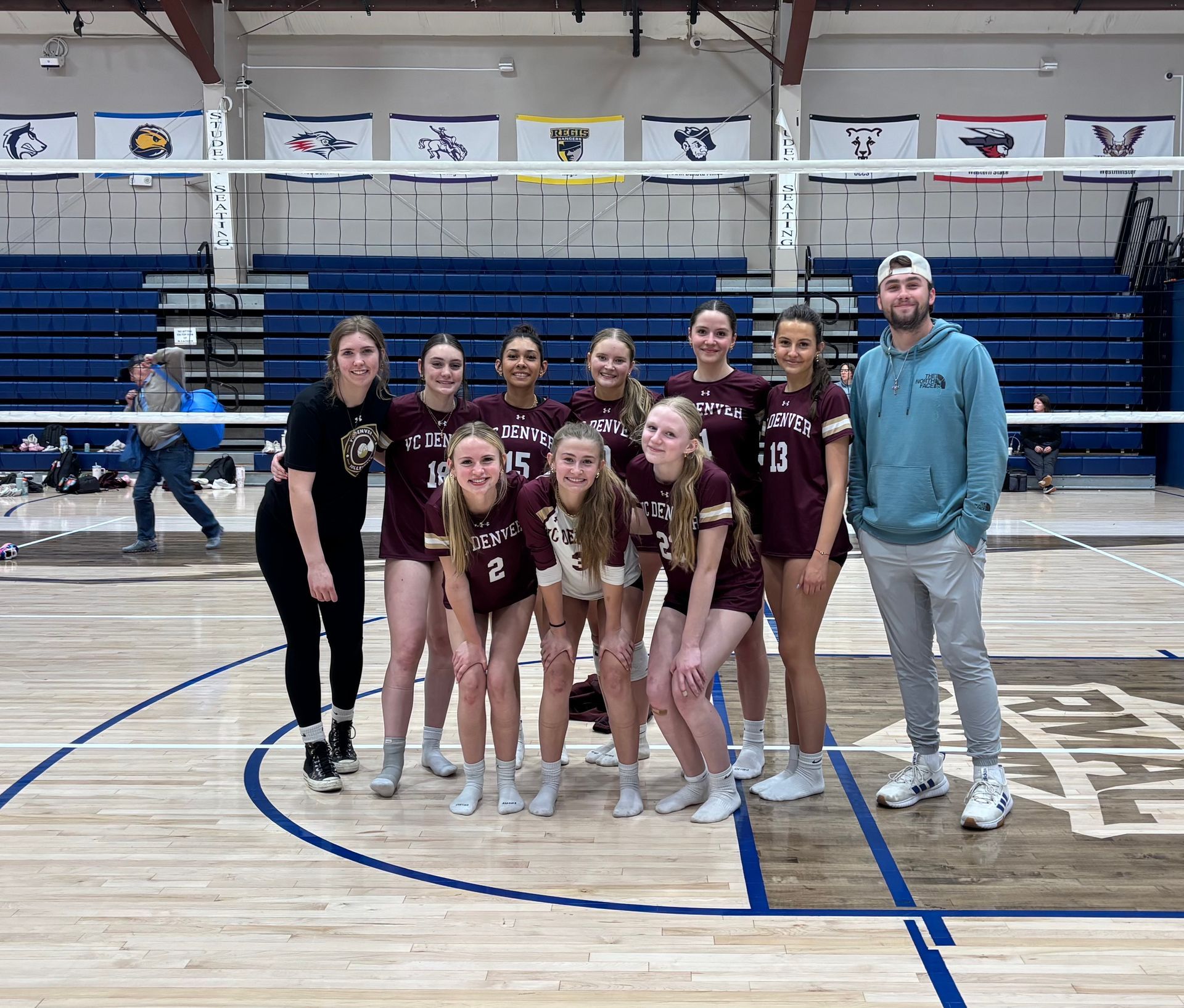 Volleyball team posing in a gym, wearing maroon uniforms with a coach standing beside them.