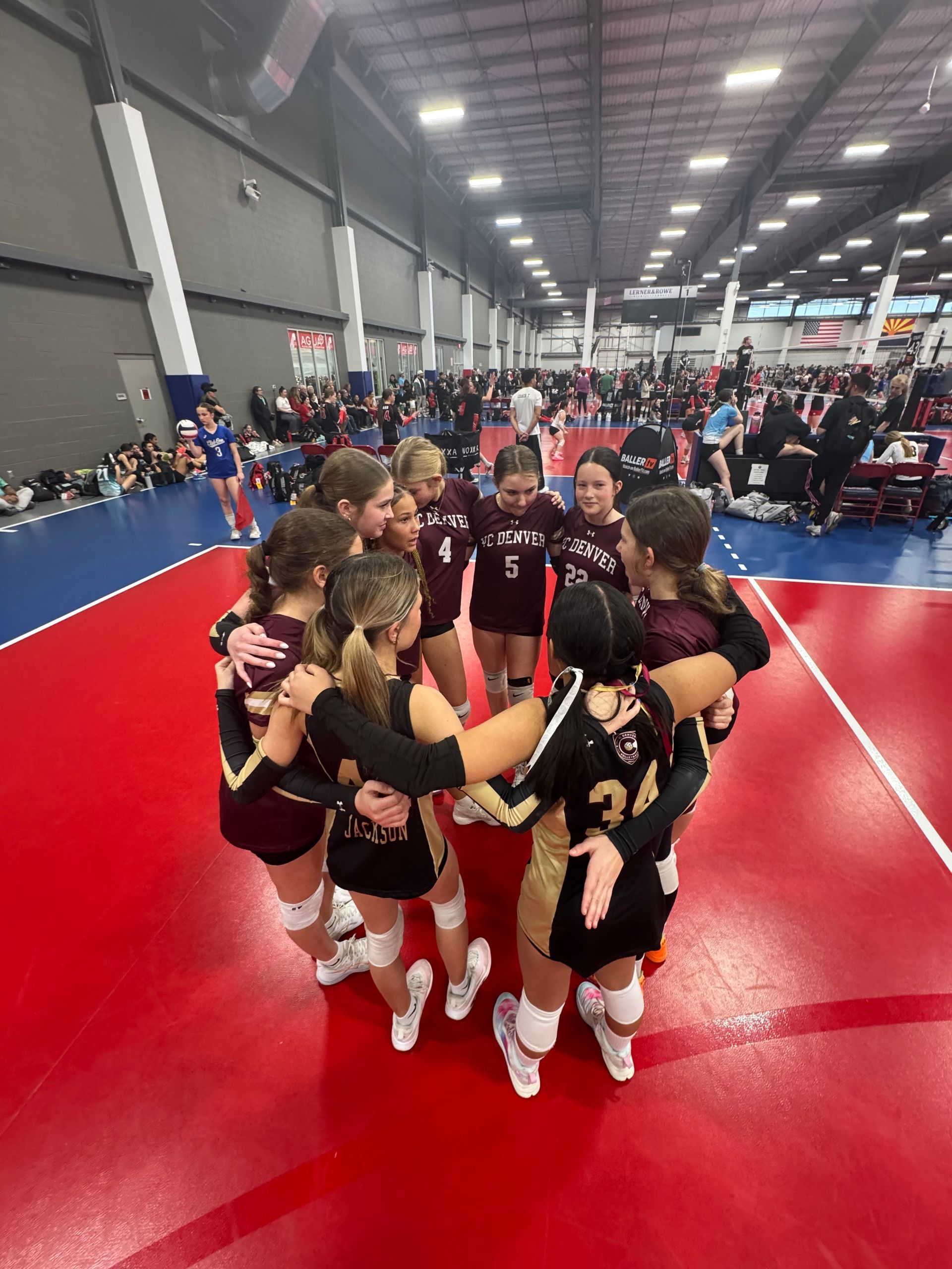 Cheer team huddles on a red gym mat inside a large indoor arena.