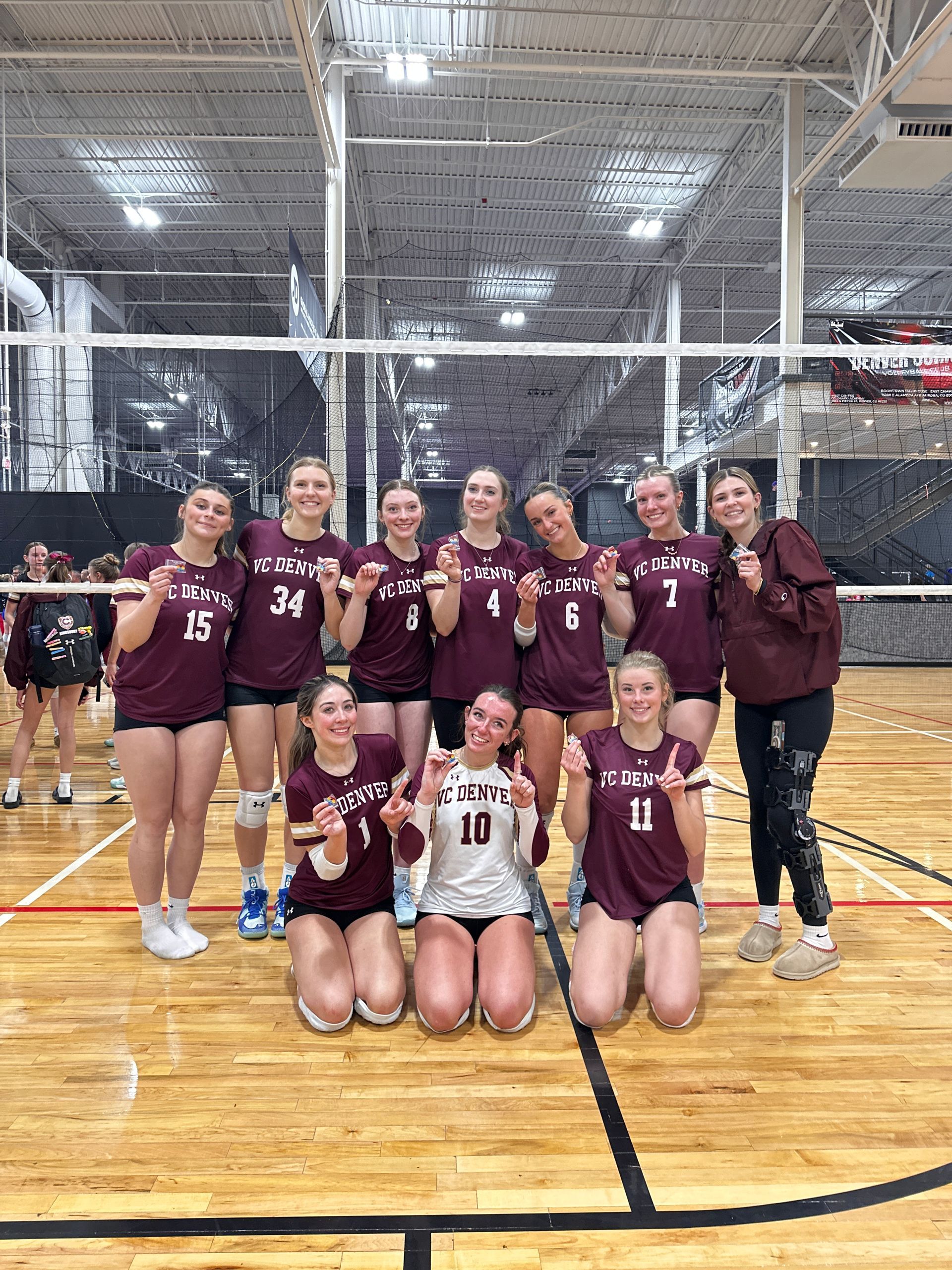 Volleyball team posing on court in maroon jerseys after a match, with net and gym visible.