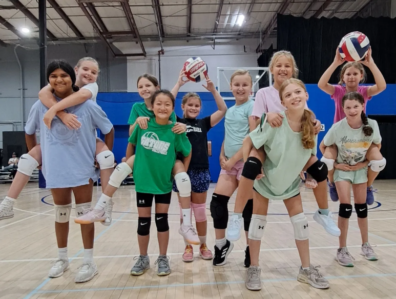 Young girls in a volleyball gym, many giving piggyback rides, holding volleyballs, smiling, and wearing knee pads.