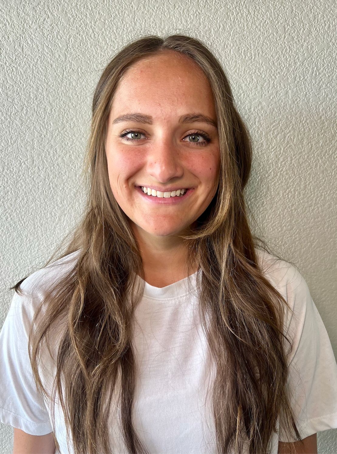Smiling person with long brown hair in a white T-shirt against a light textured wall