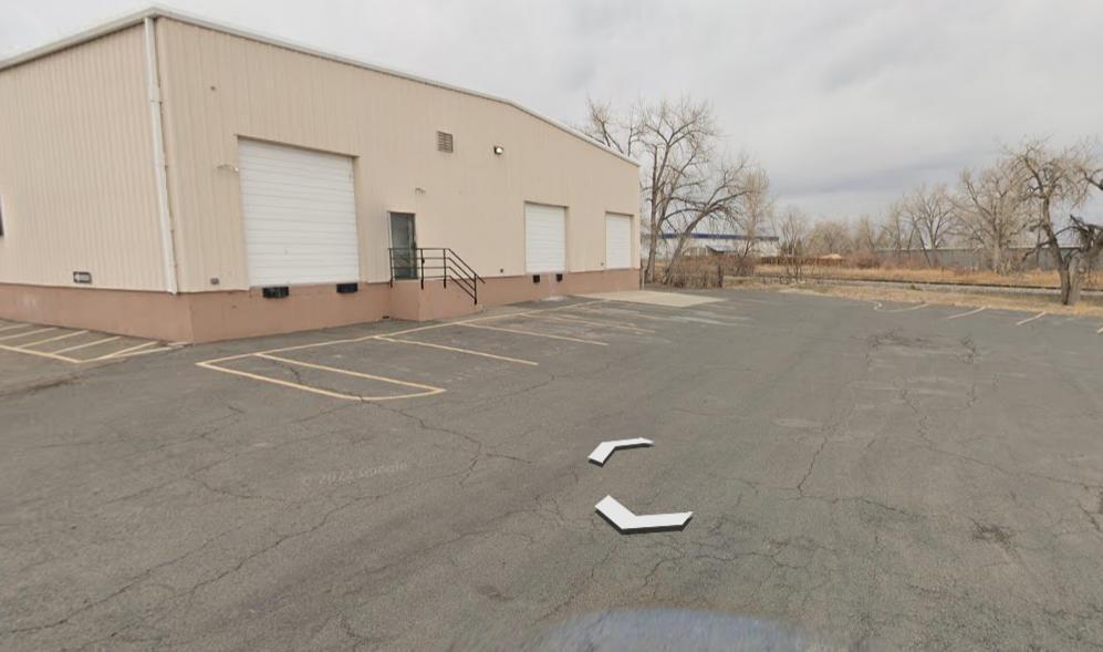 Tan industrial building with three garage doors, a paved lot, and bare trees on a cloudy day.