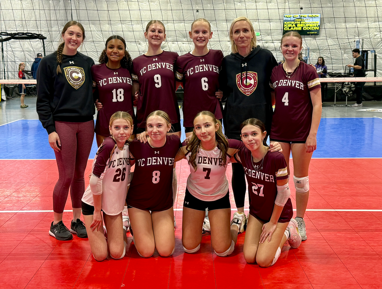 Indoor volleyball team posing on a red court, wearing maroon and black uniforms in a gym.