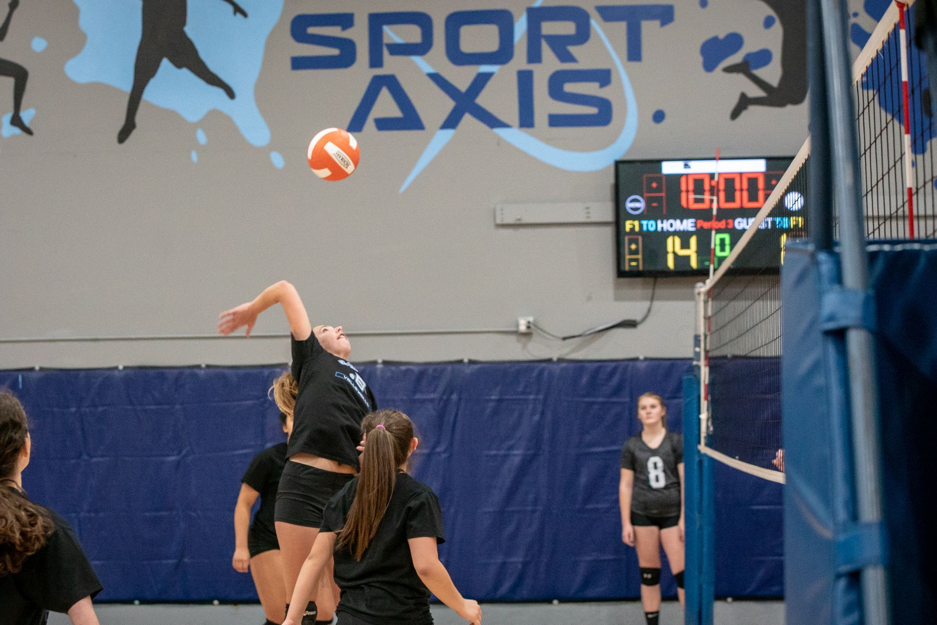 Volleyball player jumps to spike ball during game at Sport Axis; scoreboard shows the score.
