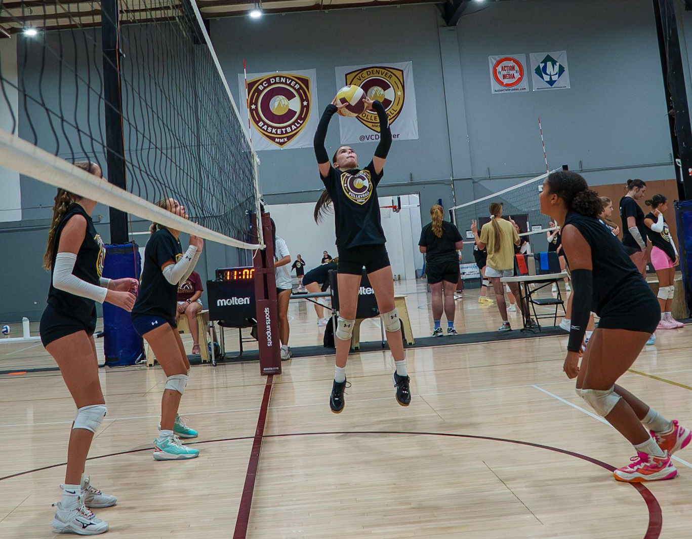 Volleyball players in a gym, with one player jumping to set the ball near the net.