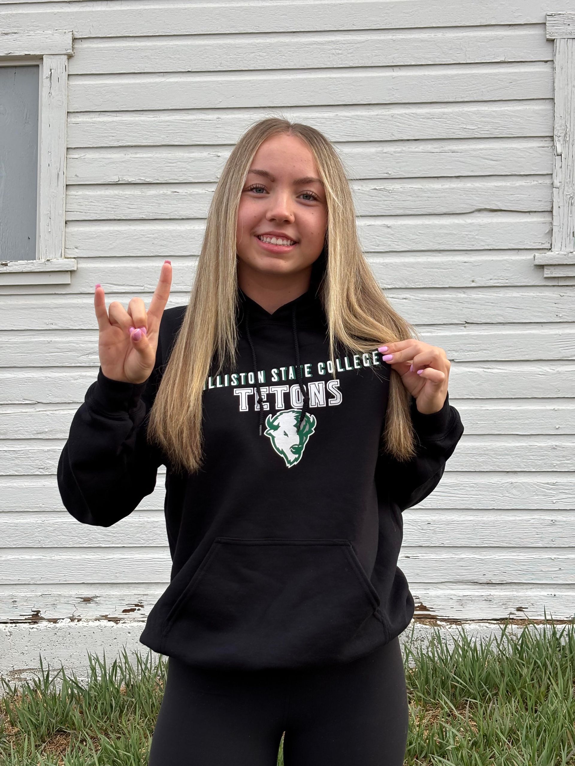 Smiling person in black hoodie and leggings posing outside a white wooden wall, making hand signs