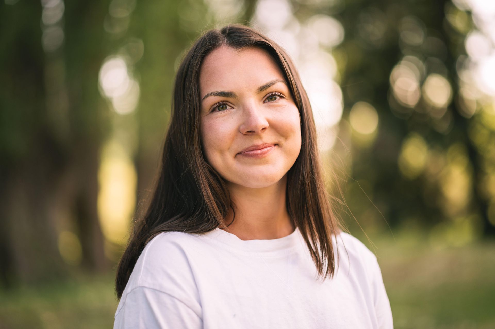 Woman with dark hair and a white shirt smiles outdoors, soft sunlight.