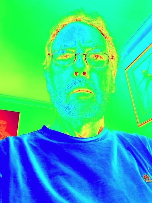 Color-inverted selfie of a person wearing glasses and a blue shirt indoors, facing the camera.