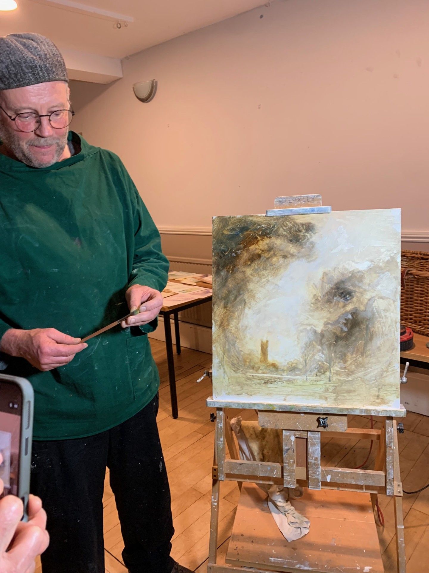 Person in green sweater standing beside a canvas on an easel, painting in a room
