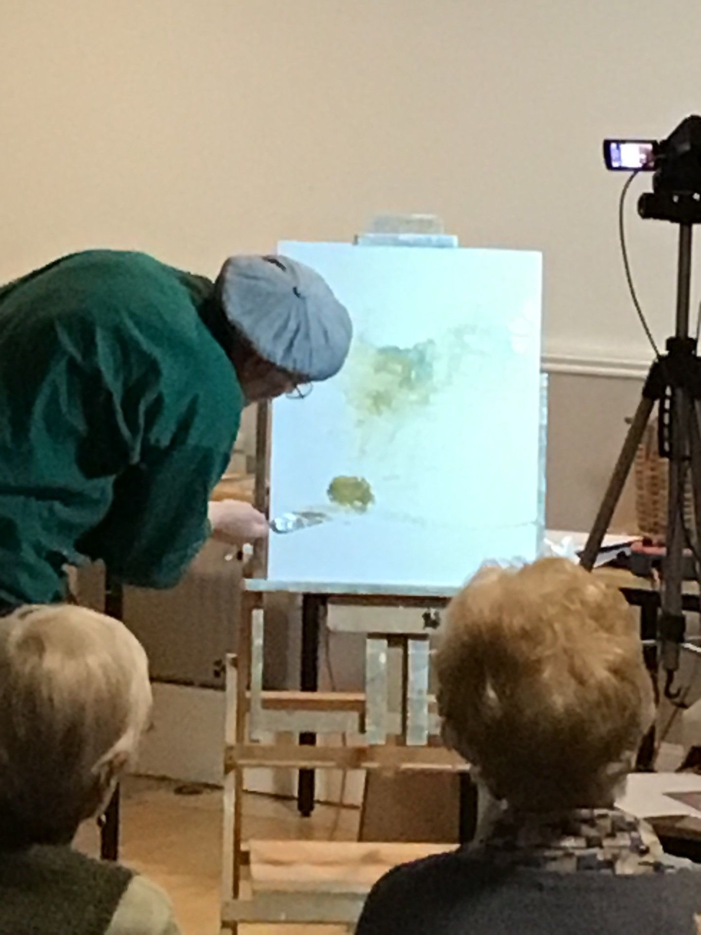 Artist painting on a canvas in front of an audience, with a camera on a tripod recording.